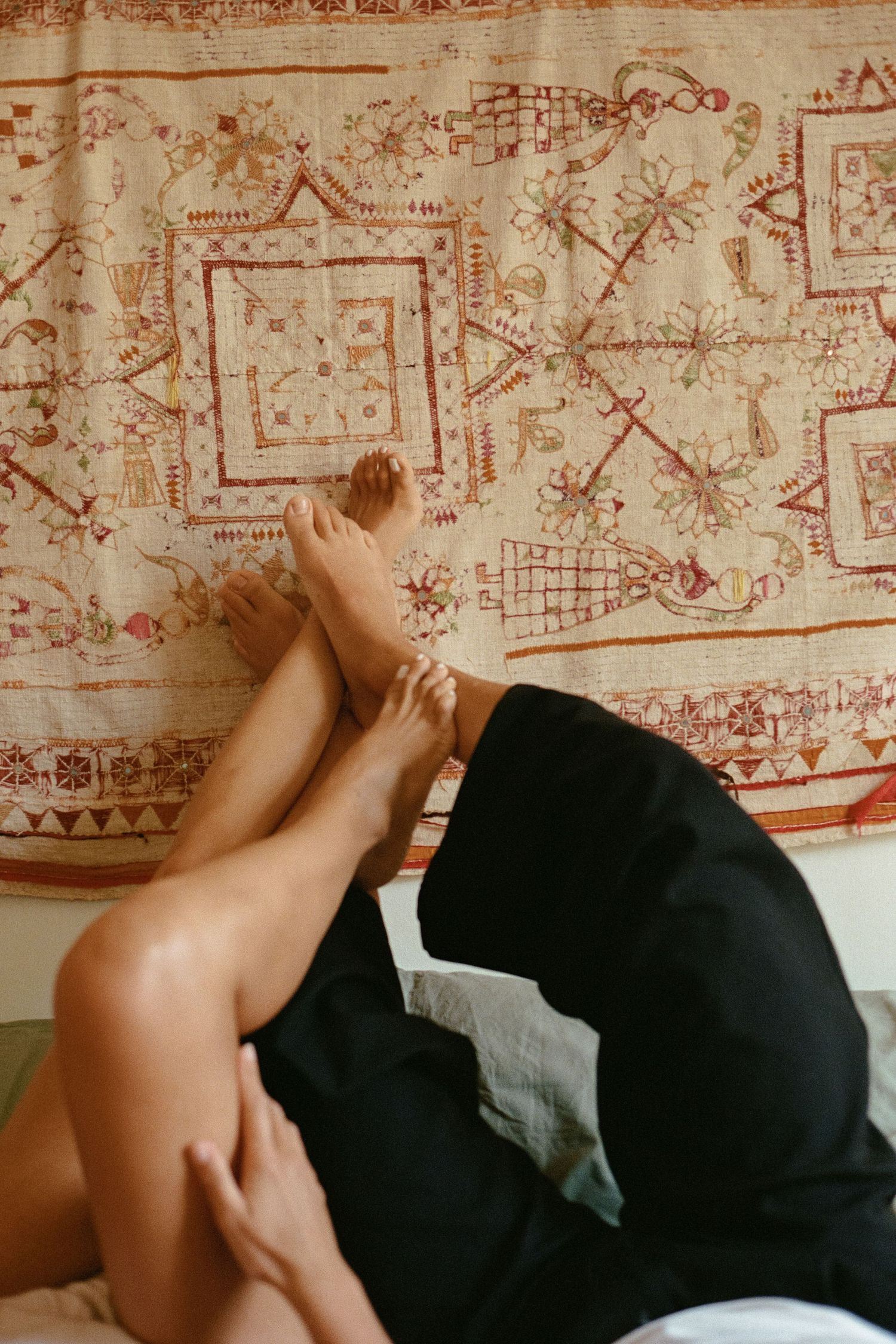Two people lie on a bed with legs raised against a decorative boho tapestry wall hanging in a cozy bedroom.