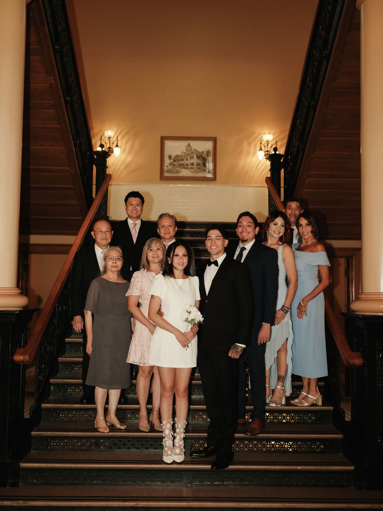 Wedding party poses on grand staircase, bride in white mini dress holds bouquet with groom in tuxedo and family.