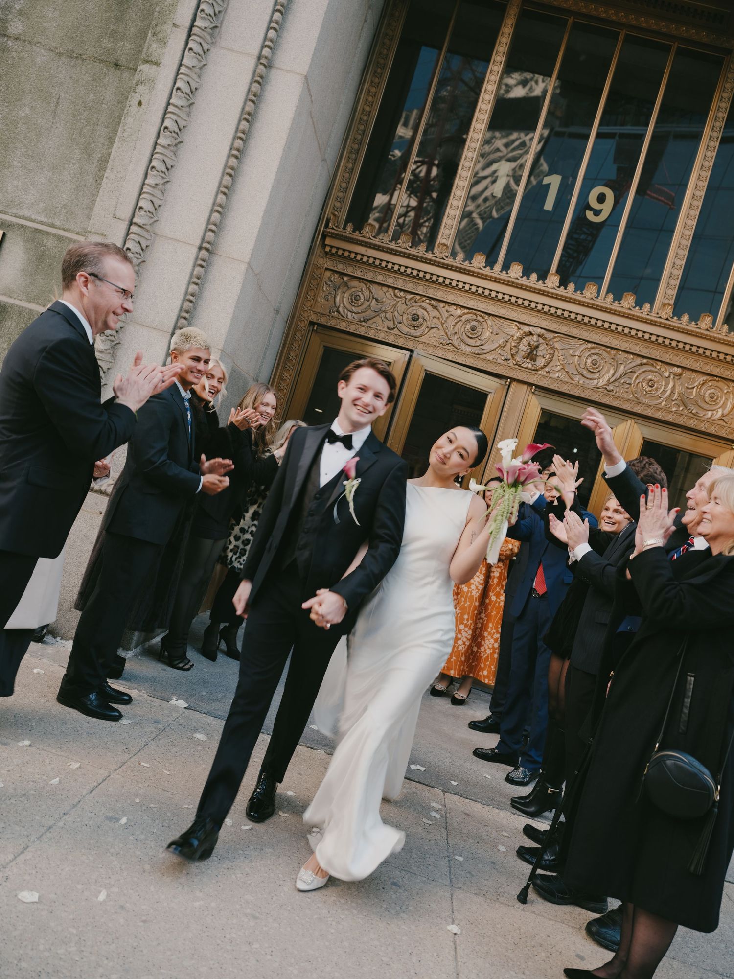 Smiling newlyweds walk through applauding guests outside ornate Art Deco building after ceremony.