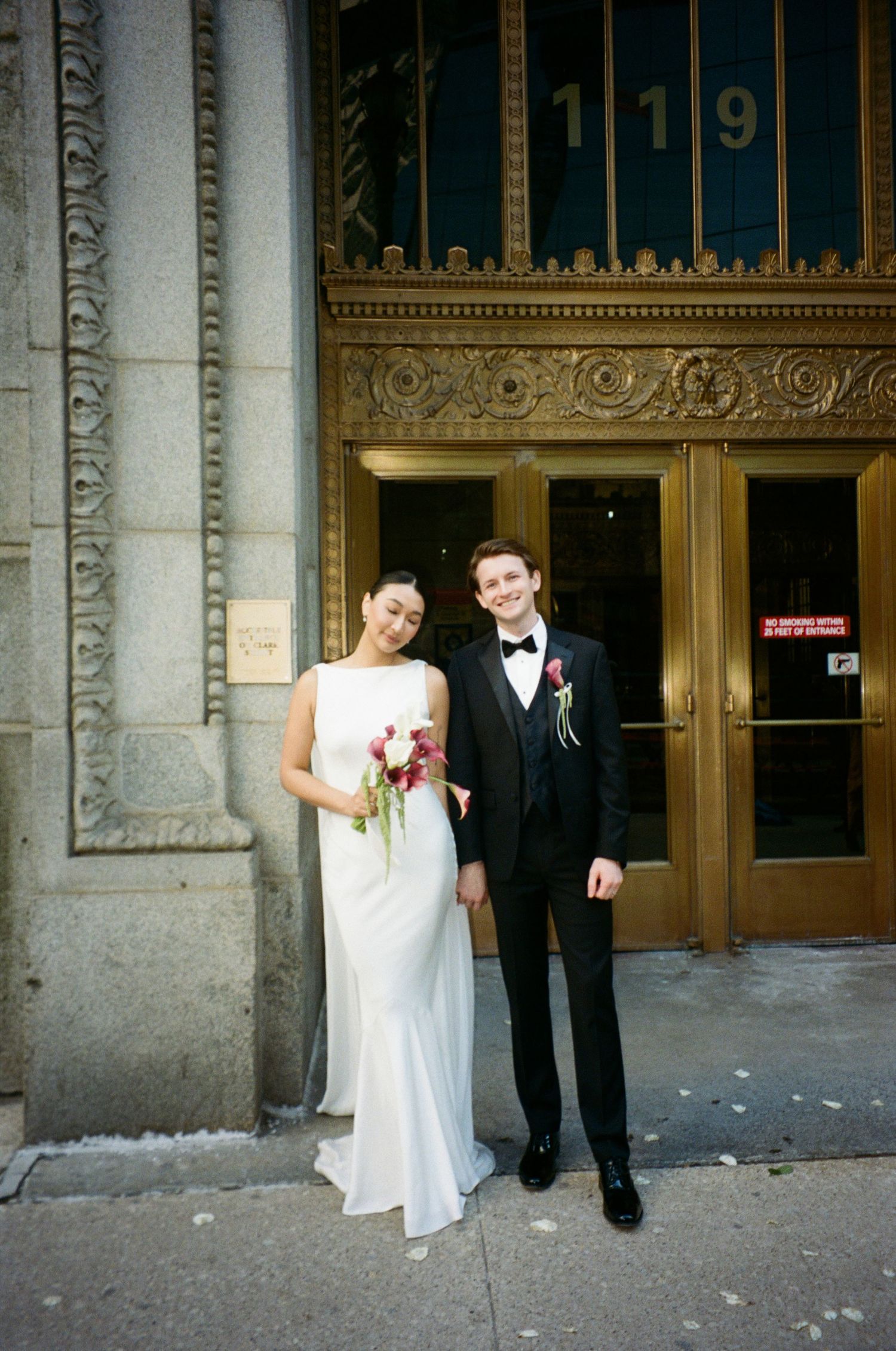 Bride and groom pose outside ornate building entrance numbered 119, bride holding red bouquet.