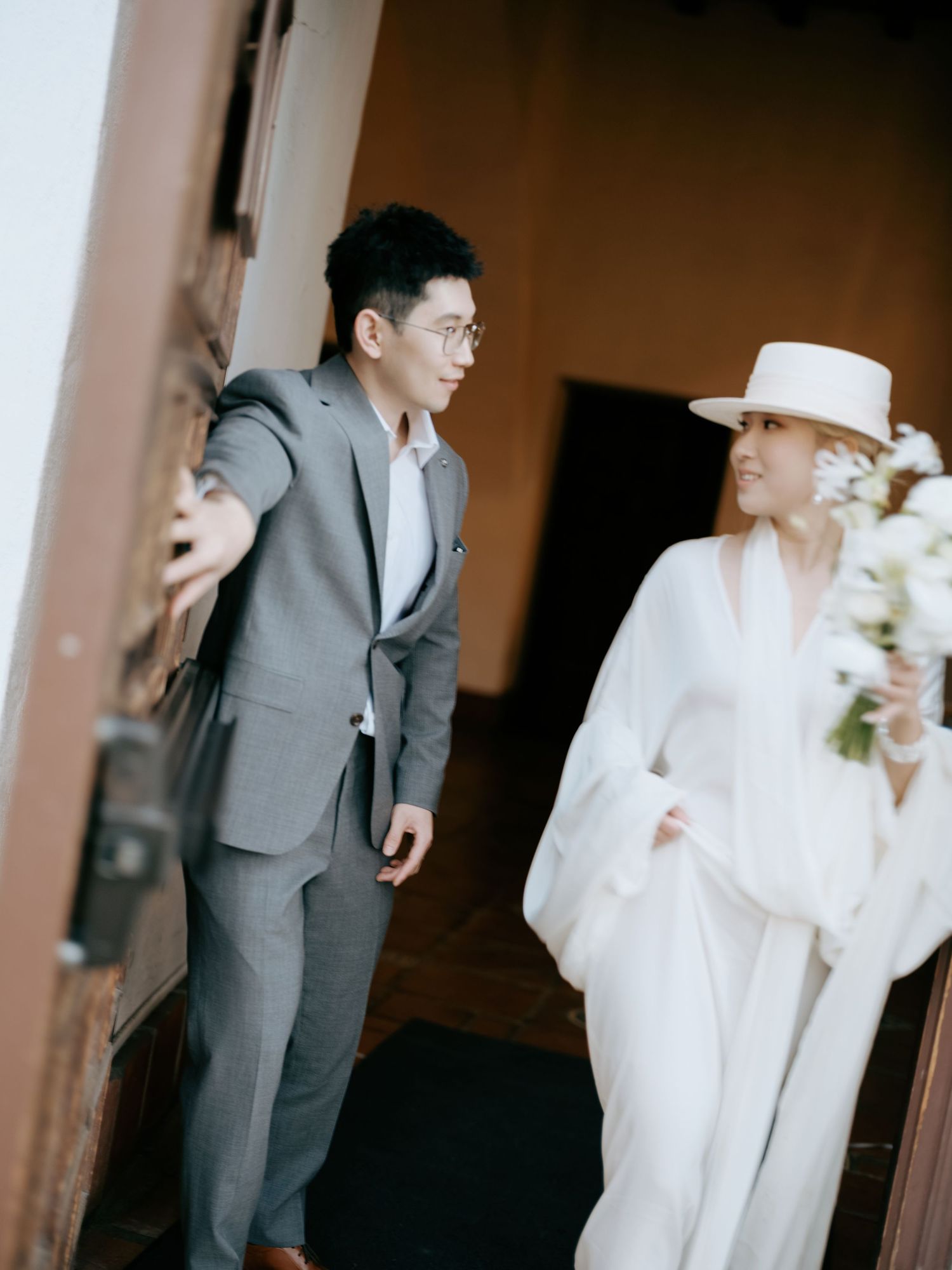 Couple sharing a glance as groom opens ornate wooden door for bride in white ensemble.