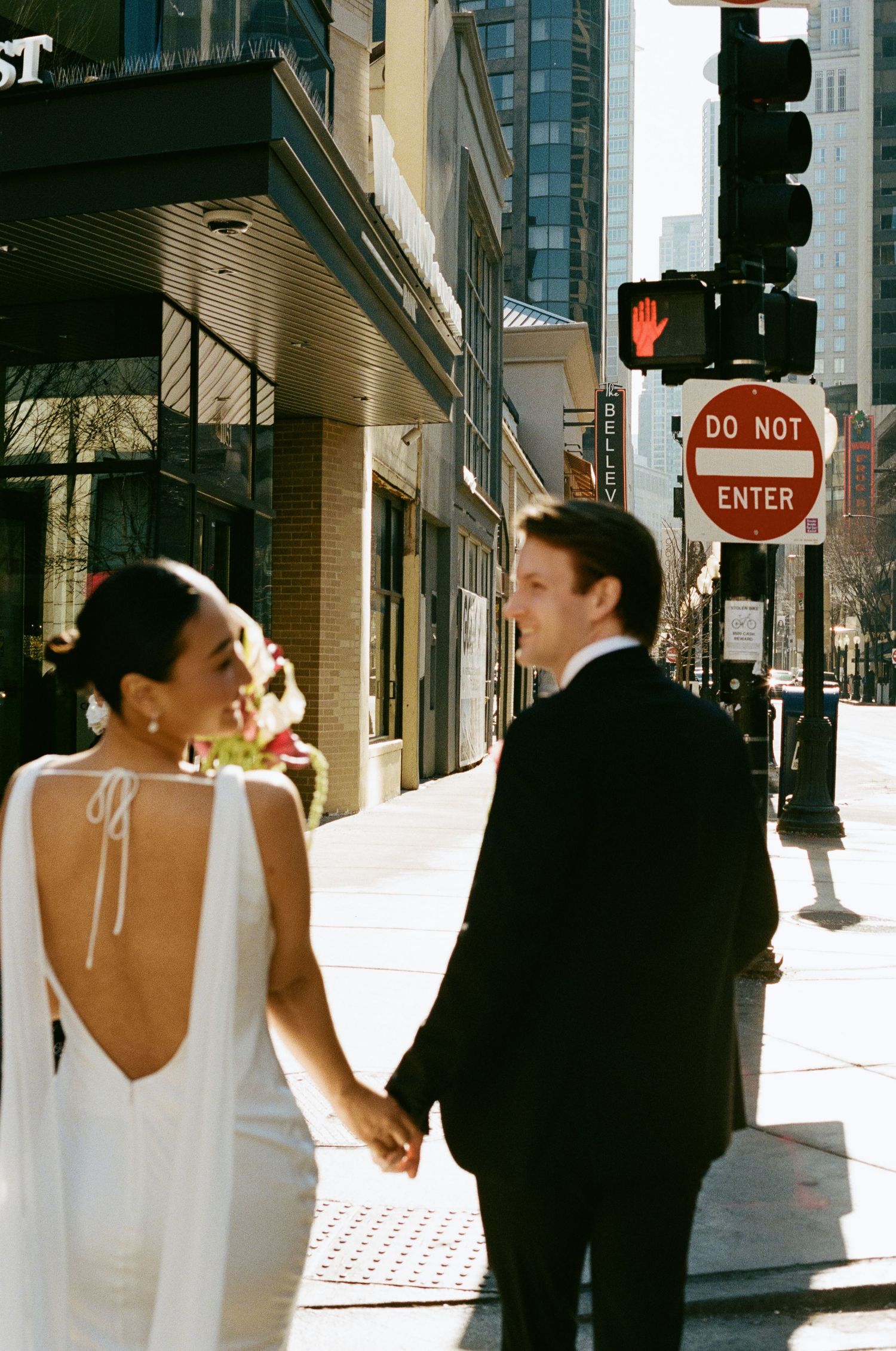 Bride in open-back white gown holds grooms hand near Do Not Enter sign on urban sidewalk.