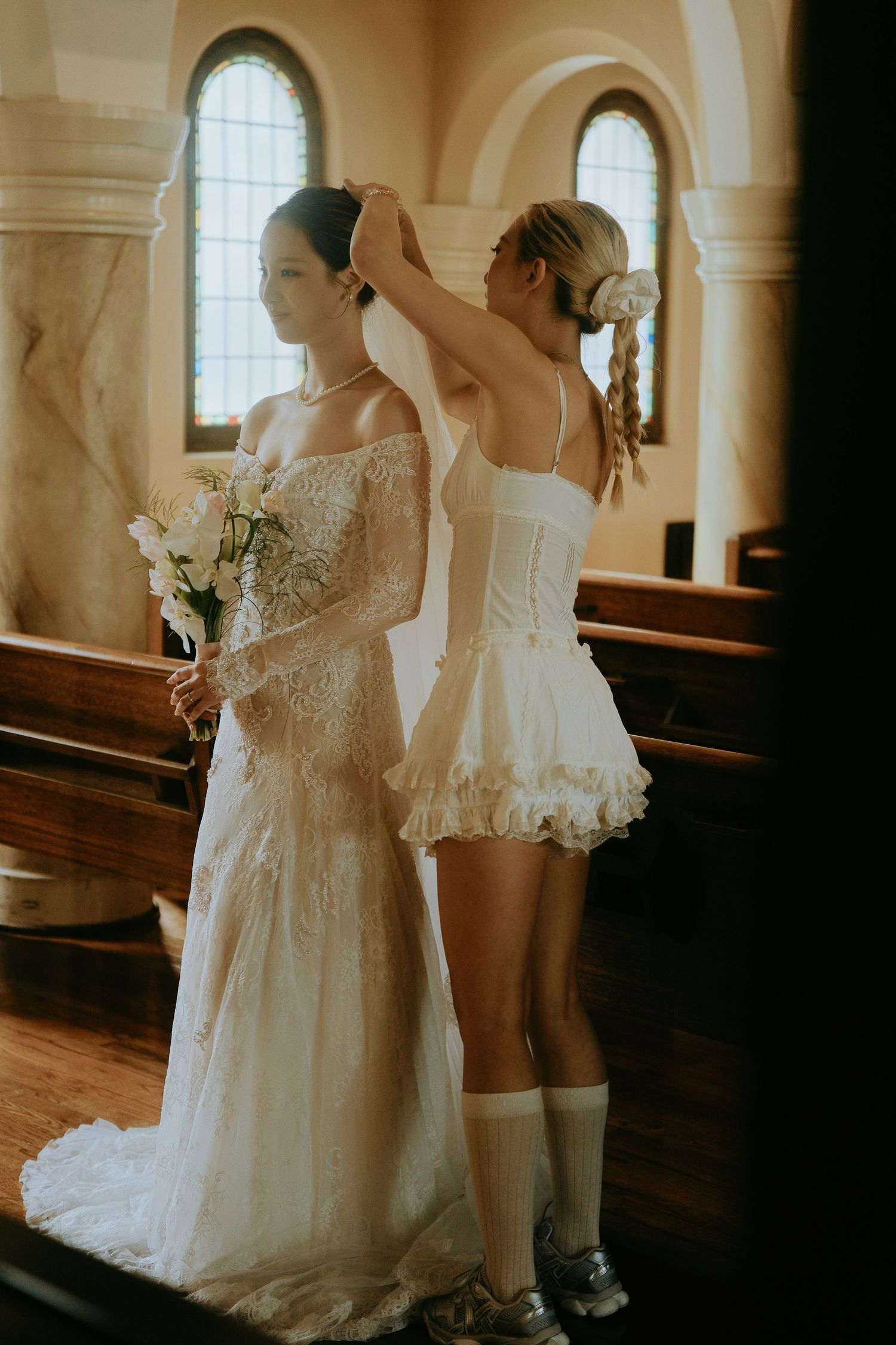 Bridesmaid in white corset adjusts brides veil inside rustic church with arched windows.