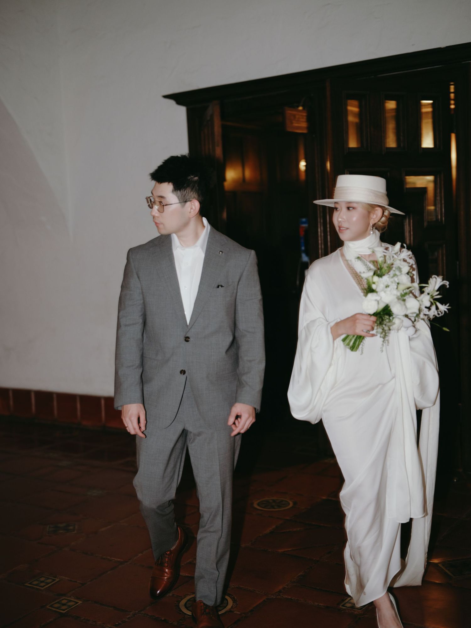 Smiling bride in white cape and hat walks alongside groom in gray suit holding bouquet.