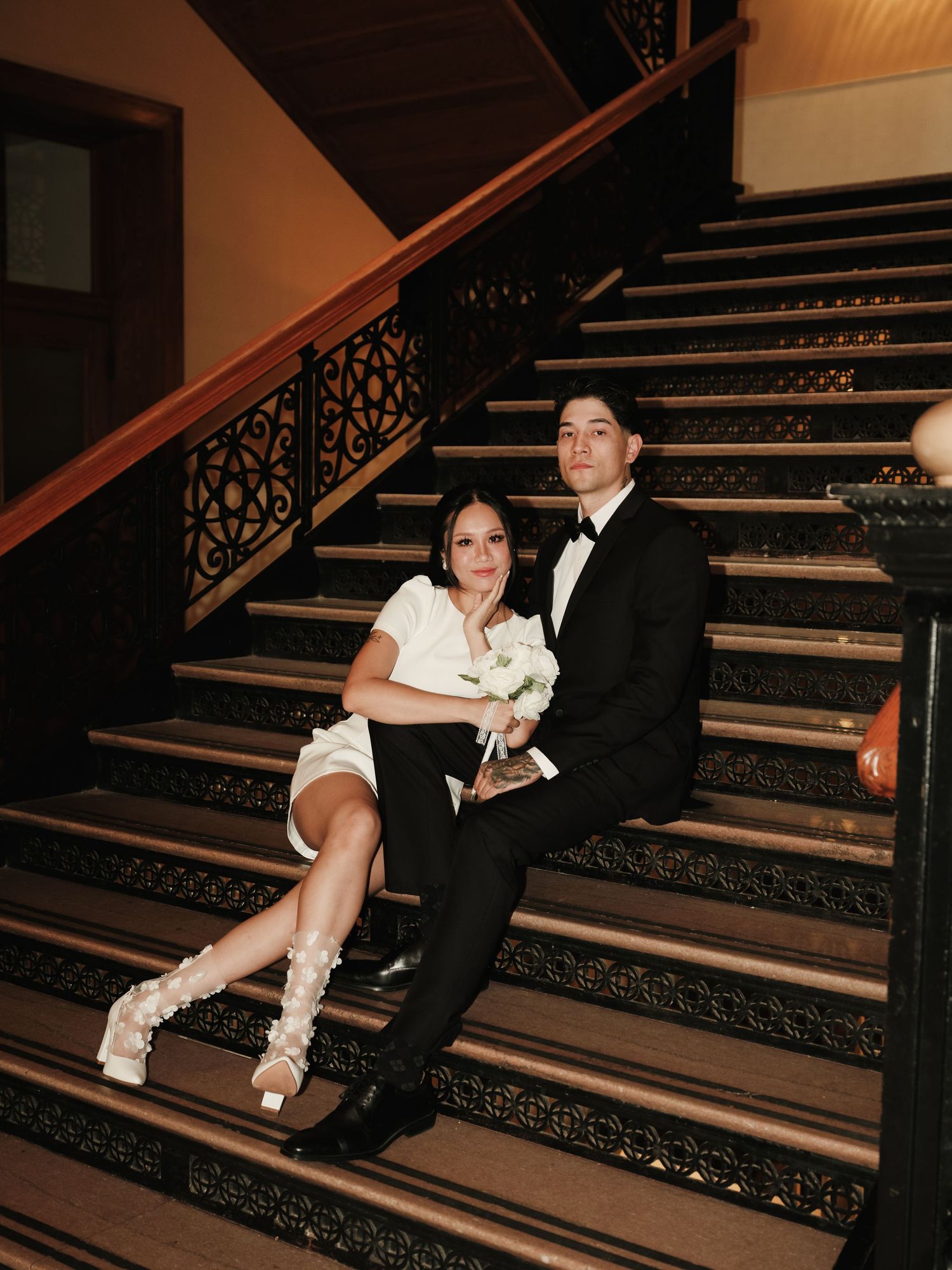 Elegant newlyweds pose together on ornate dark wooden staircase with white bridal bouquet.