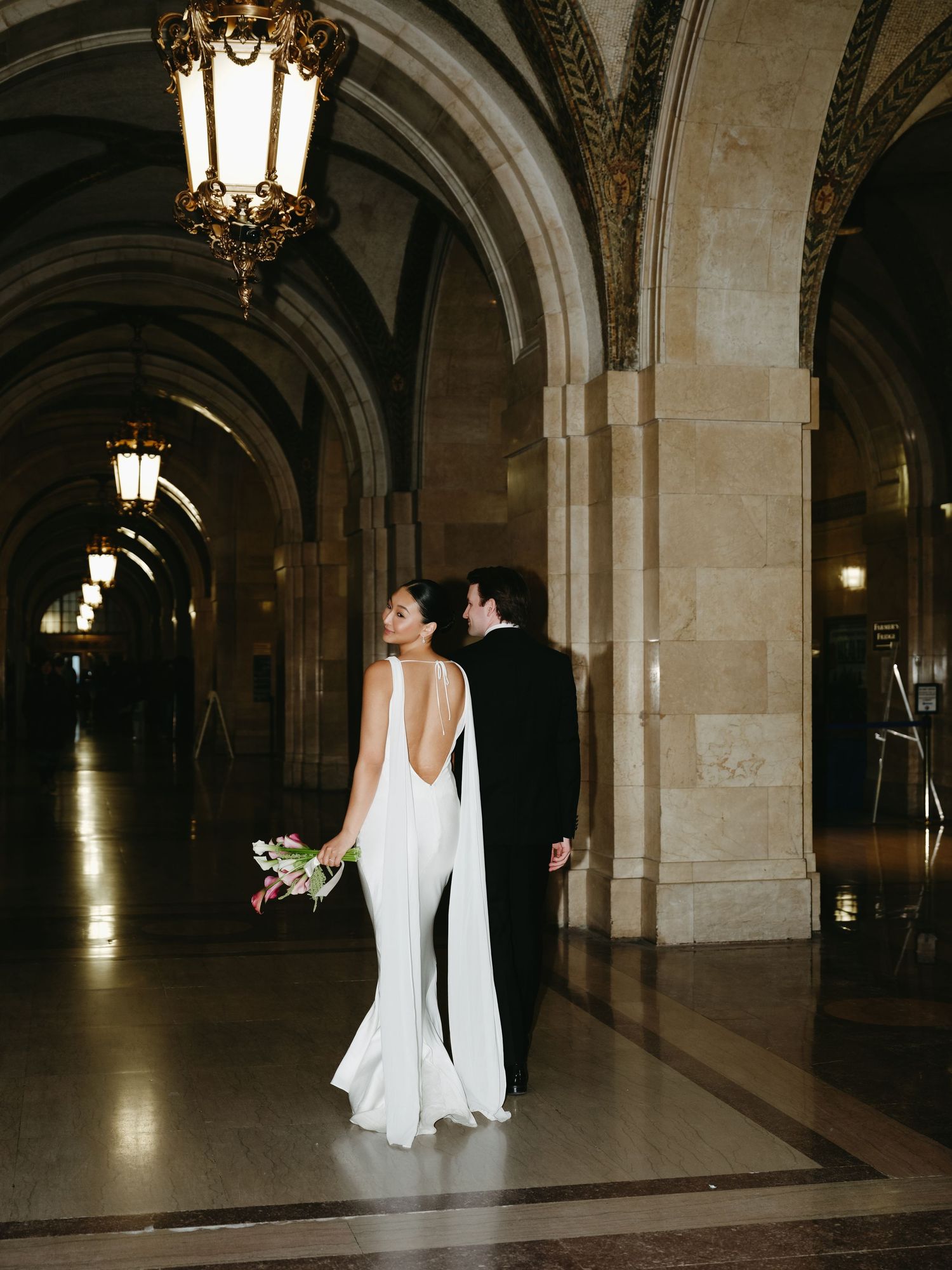 Bride in stunning backless white cape gown walking with groom through grand arched corridor.