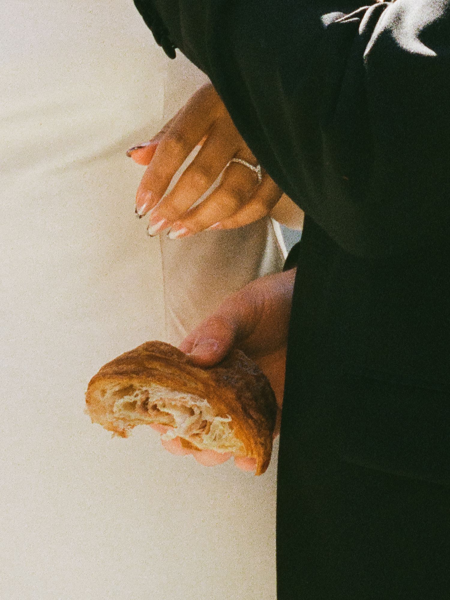 Close-up of bride holding a bitten croissant with silver rings beside groom in black suit.