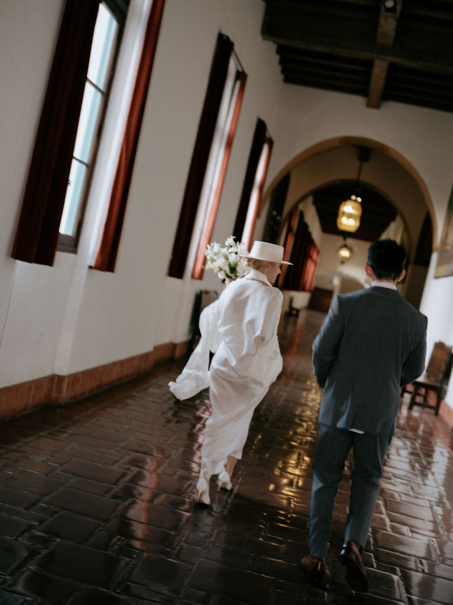 Bride in white cape and hat leads groom down tiled corridor of historic Spanish venue.