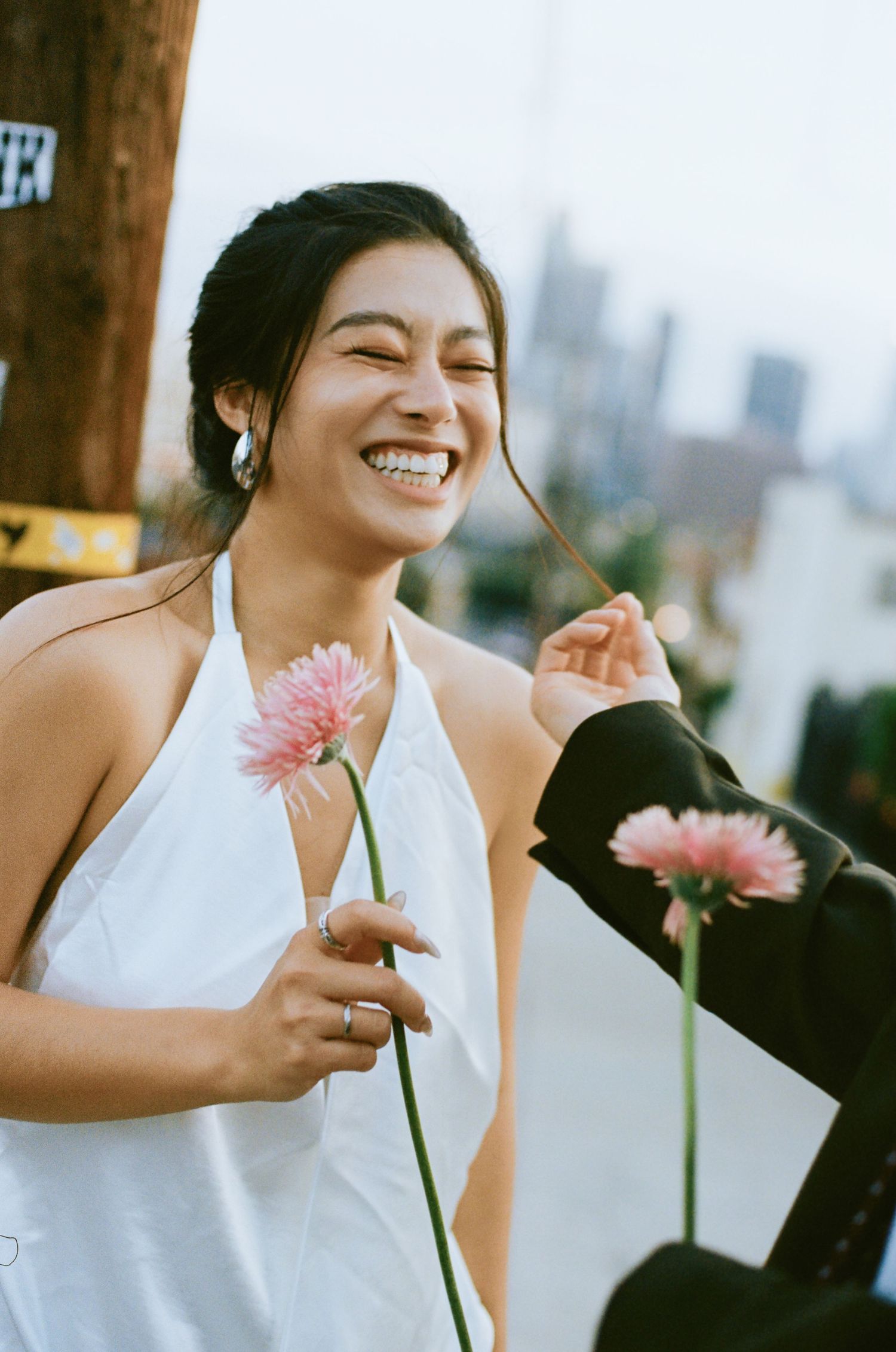 Laughing bride in white halter dress holds pink gerbera daisy with city skyline visible in background.