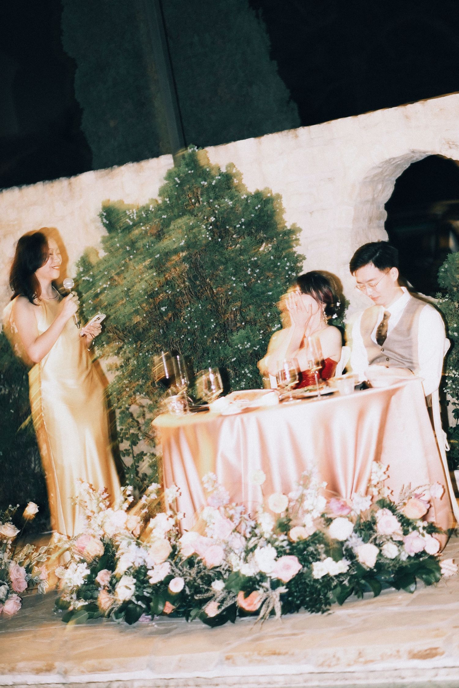 Bride and groom seated at sweetheart table with floral arrangements while bridesmaid gives speech at elegant wedding reception.