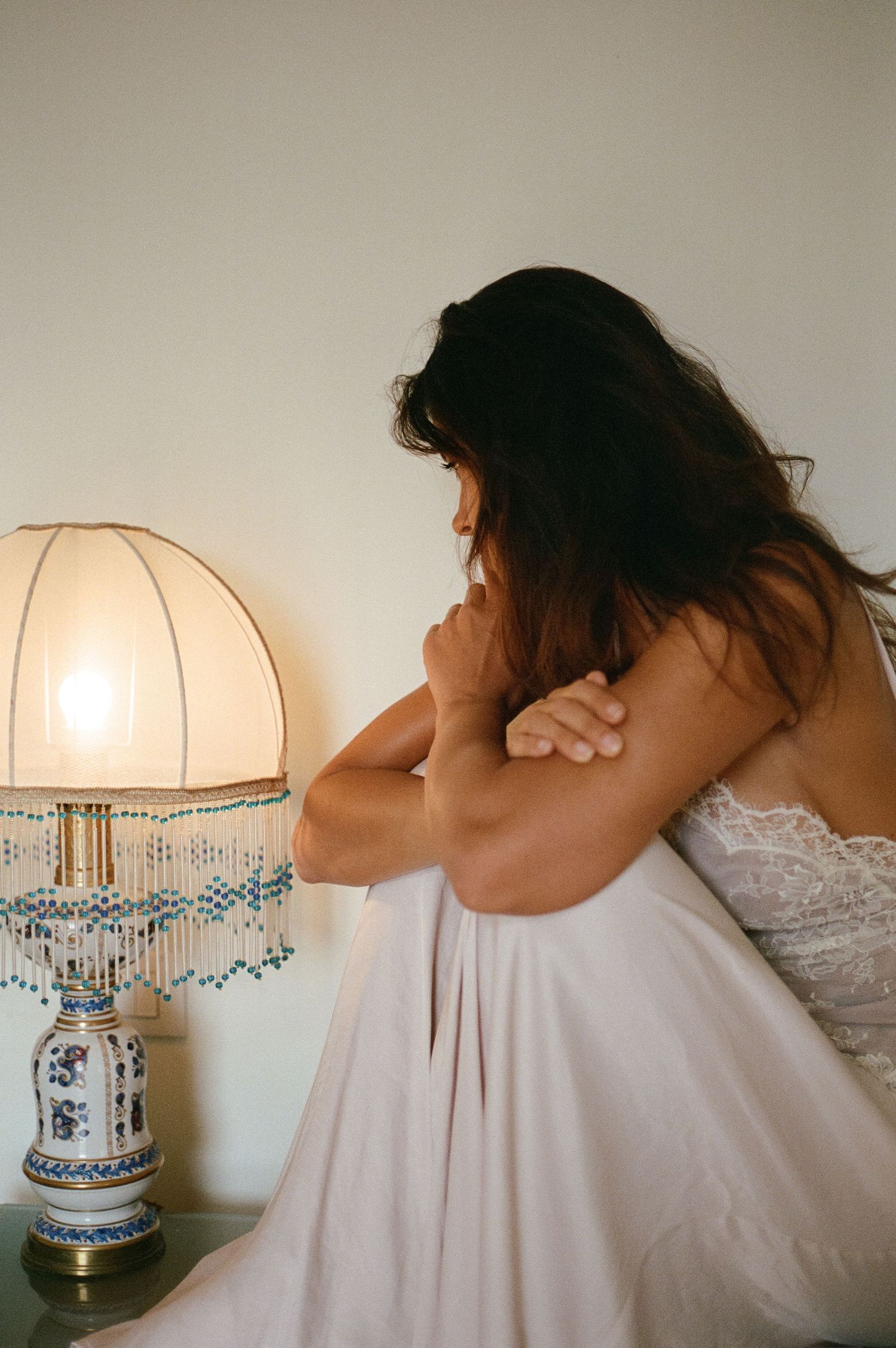 Bride in white lace gown sitting pensively beside a vintage beaded table lamp.
