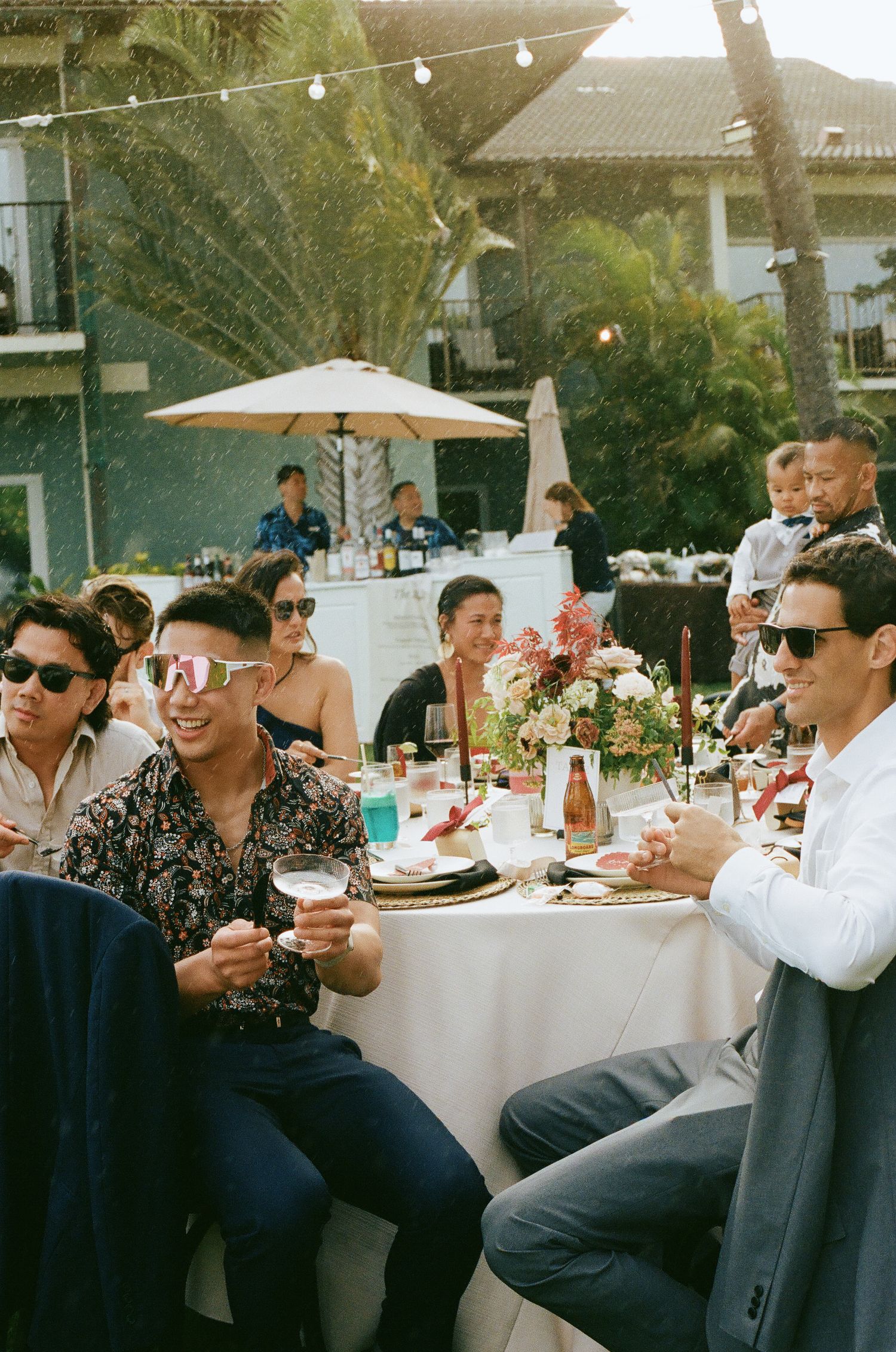 Guests in sunglasses celebrating at an outdoor wedding reception dinner table with floral decor.