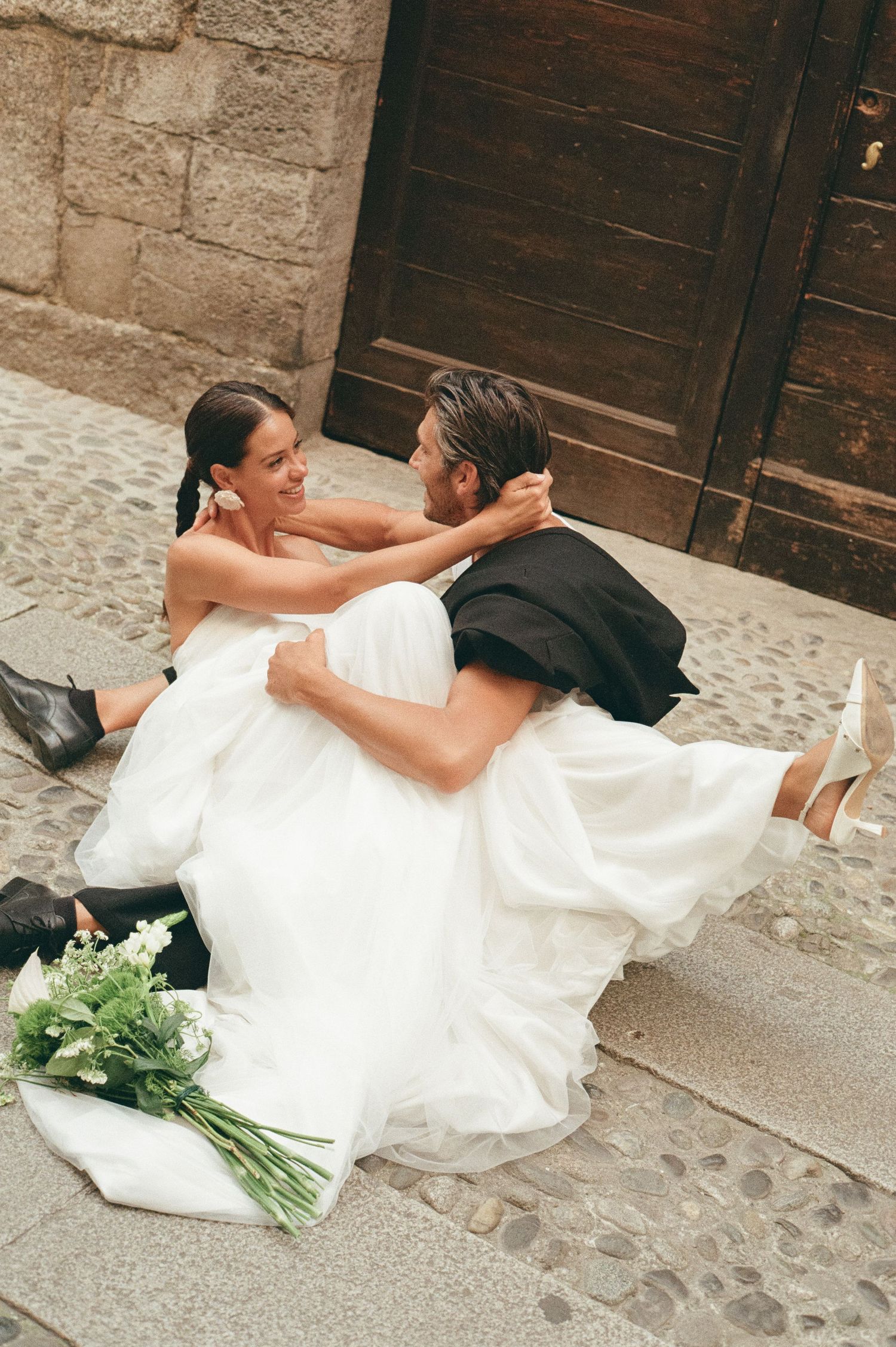 Groom holding bride in his arms while sitting on cobblestone steps outside a rustic wooden door.