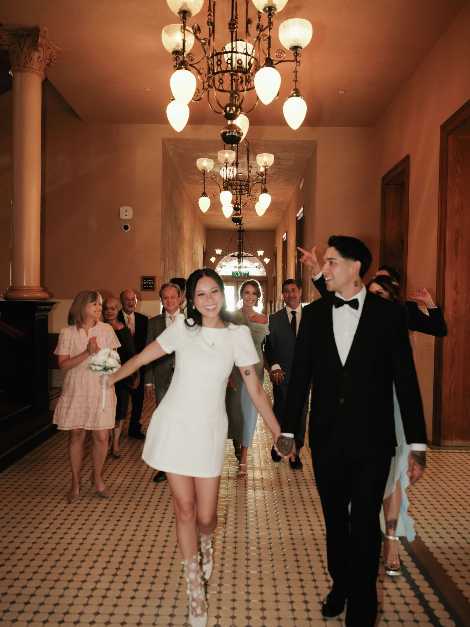 Joyful newlyweds walk hand-in-hand down ornate hallway with chandeliers as guests celebrate behind them.