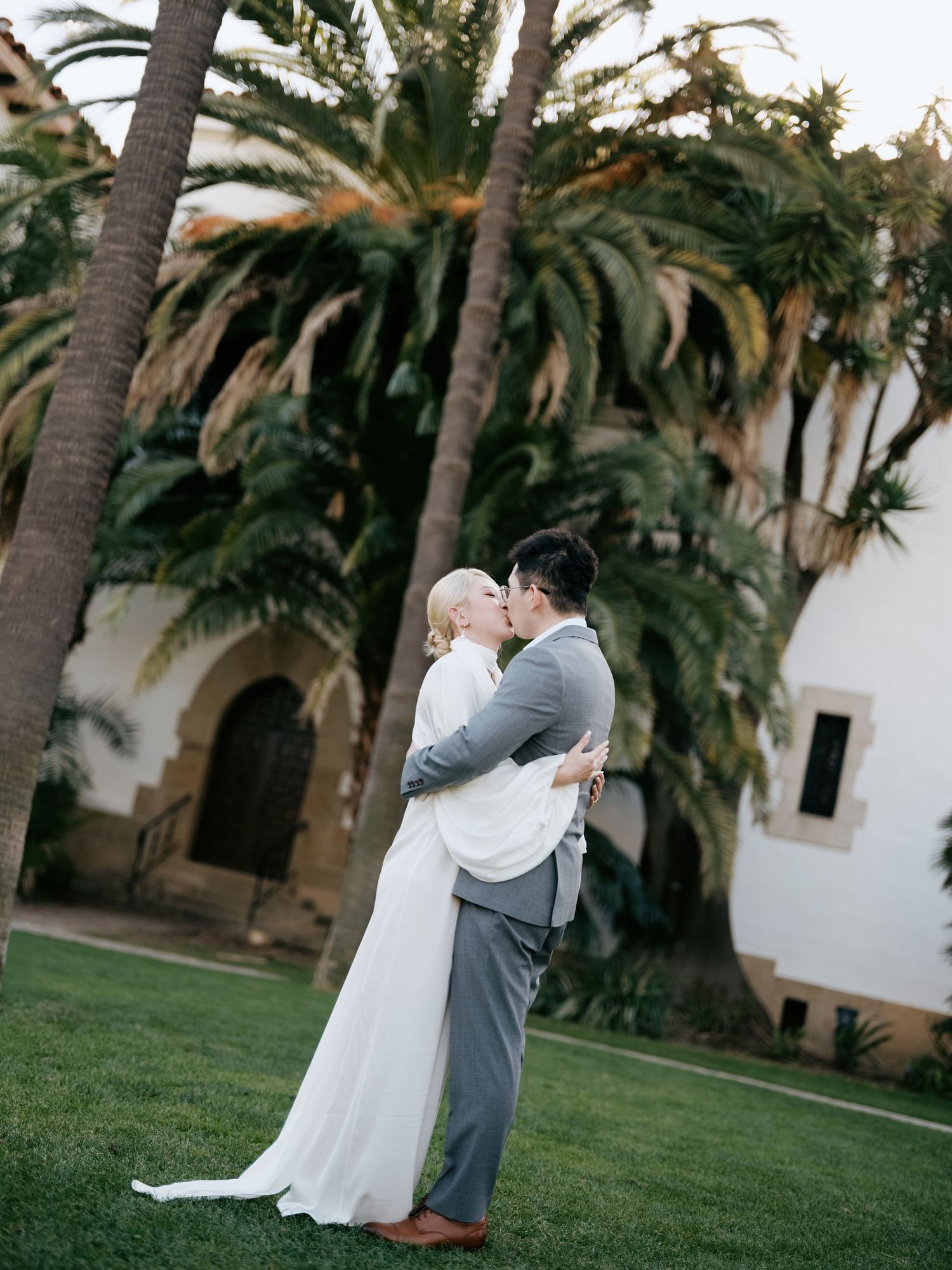 Bride and groom share first kiss on lush green lawn surrounded by tall palm trees.