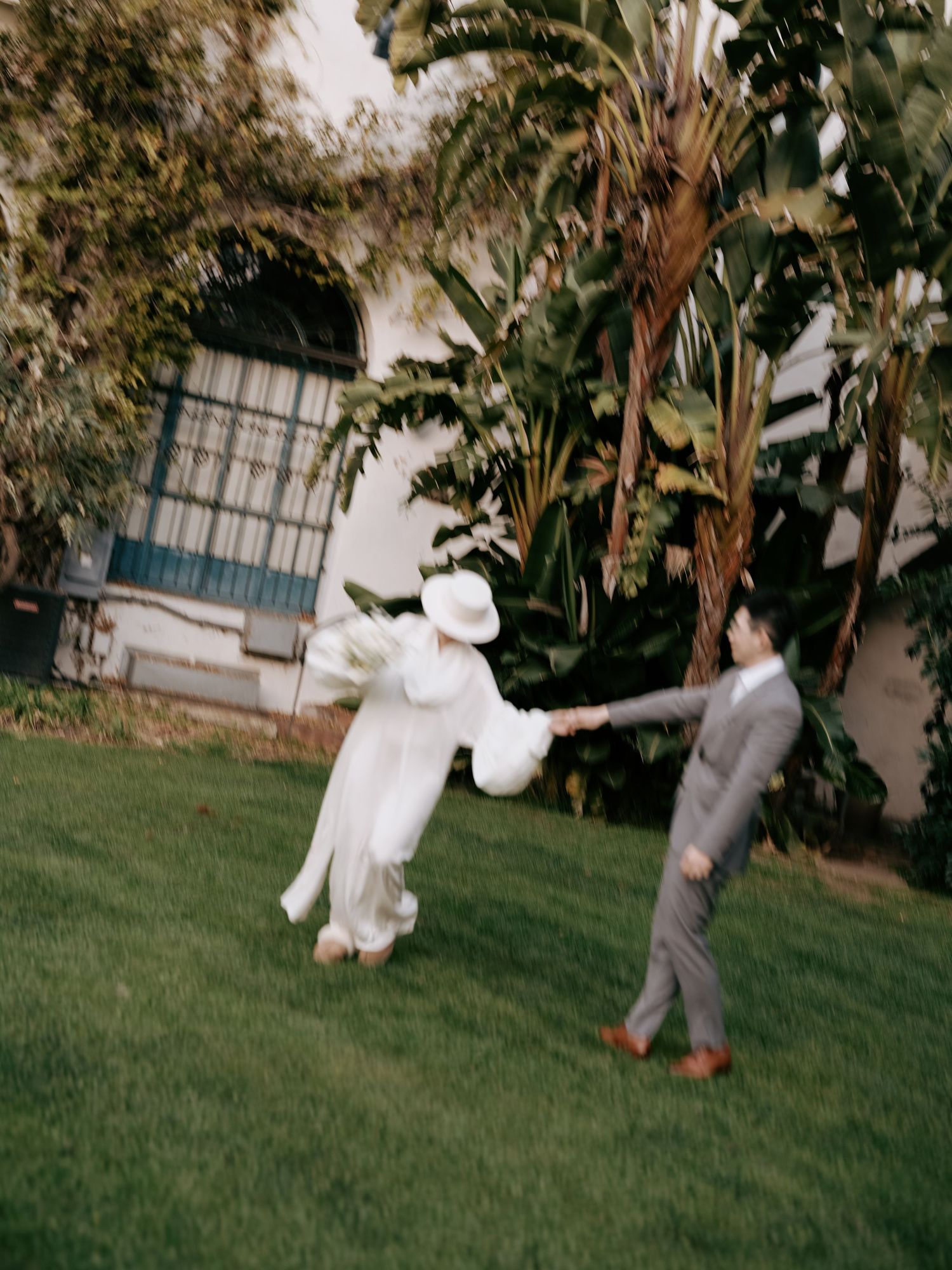 Joyful bride in white cape twirls while holding groom's hand on lush garden lawn.