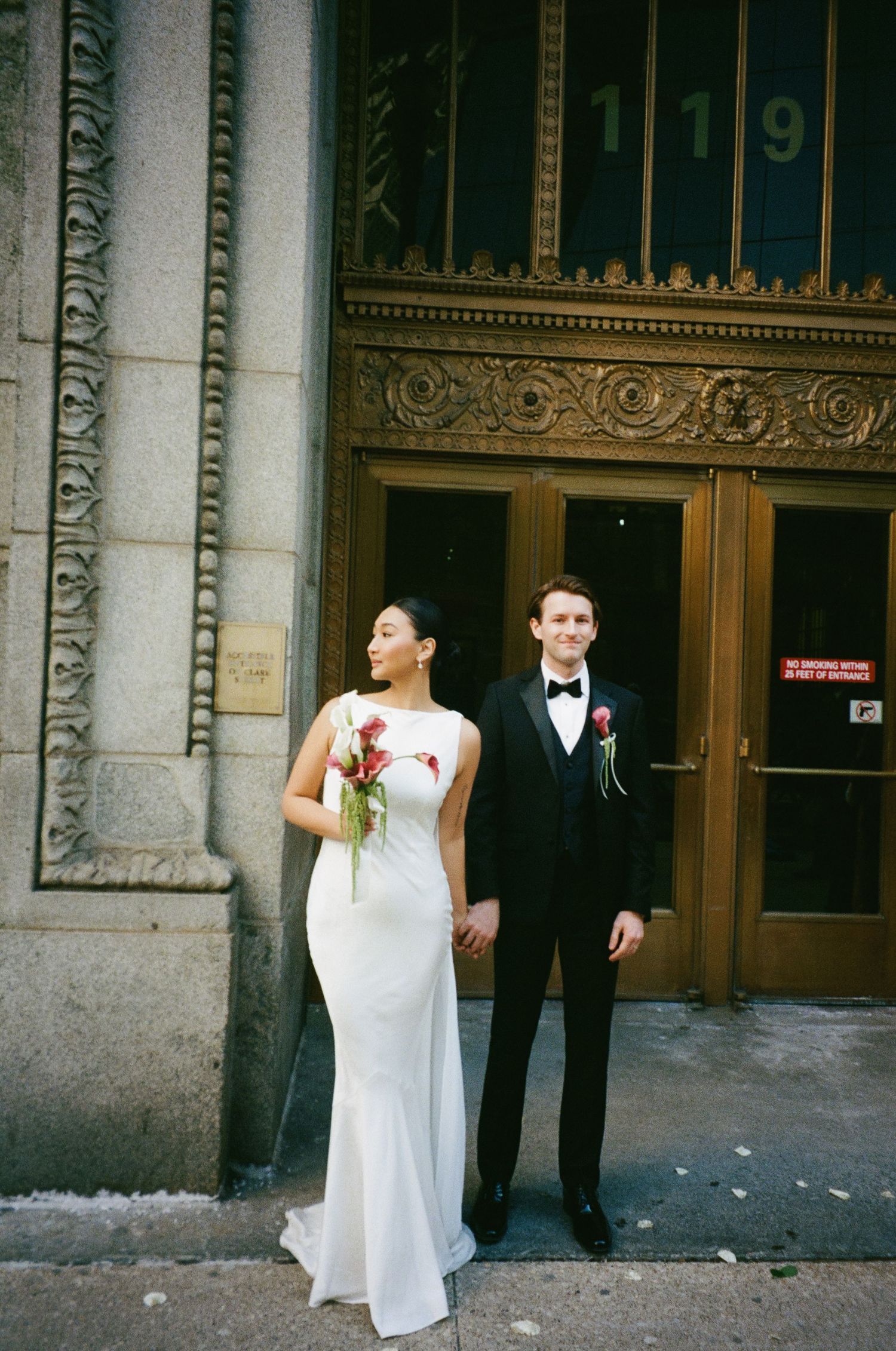 Bride and groom stand before gold-adorned building entrance 119, holding hands with red floral bouquet.