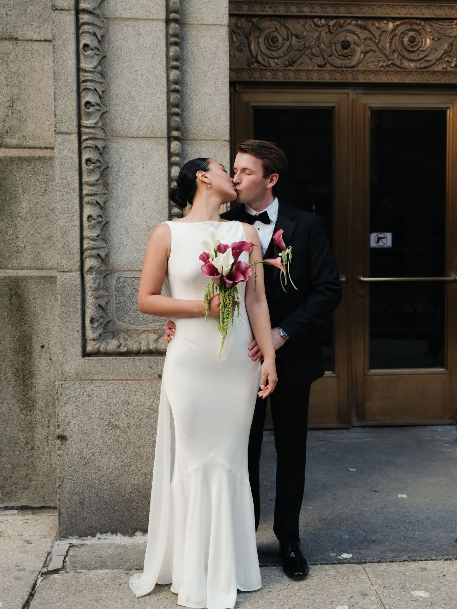 Groom kisses bride beside ornate stone building exterior, bride holding pink calla lily bouquet.