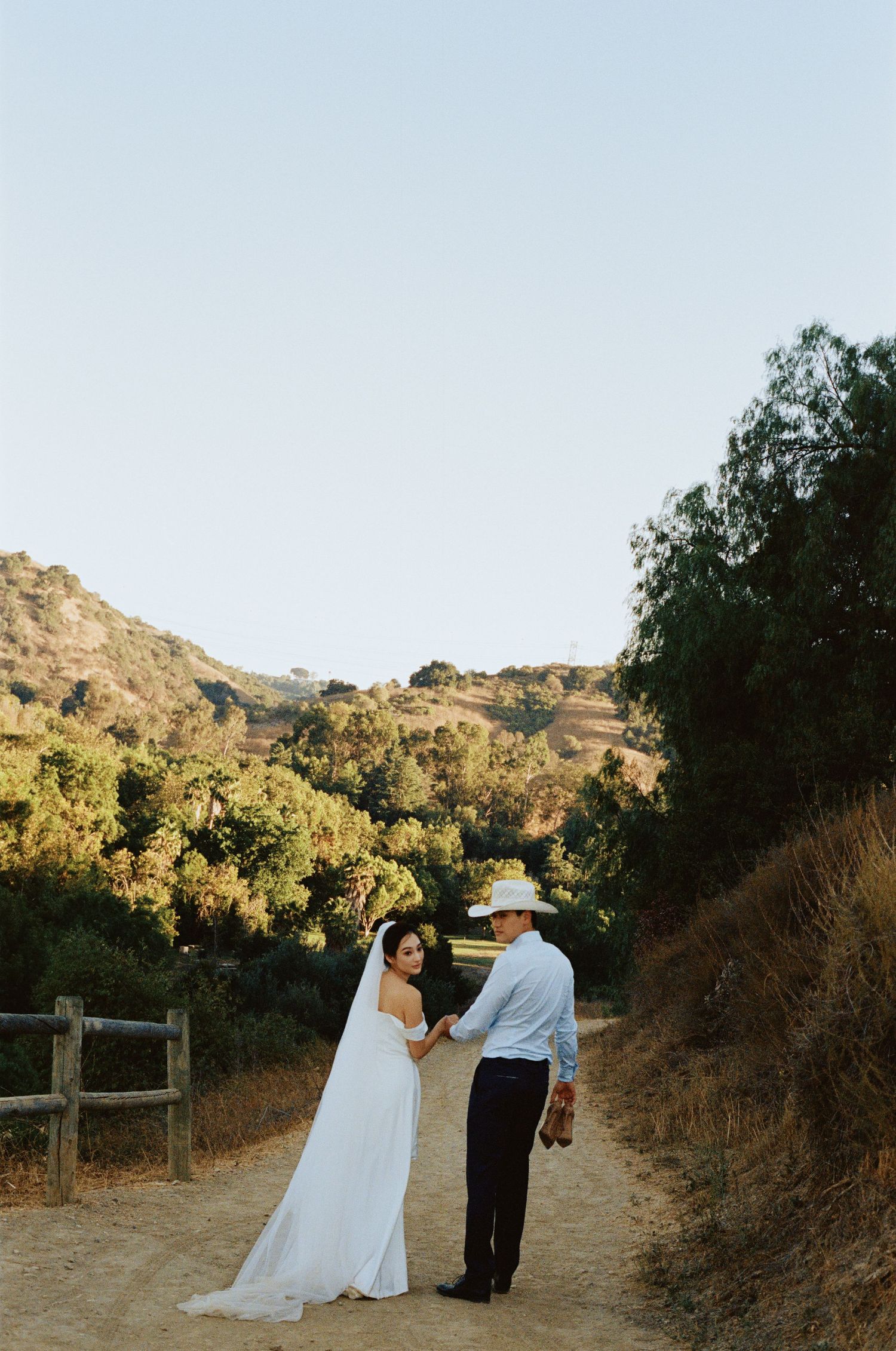 Bride and groom hold hands on a scenic dirt path surrounded by golden hills and lush greenery during outdoor wedding.