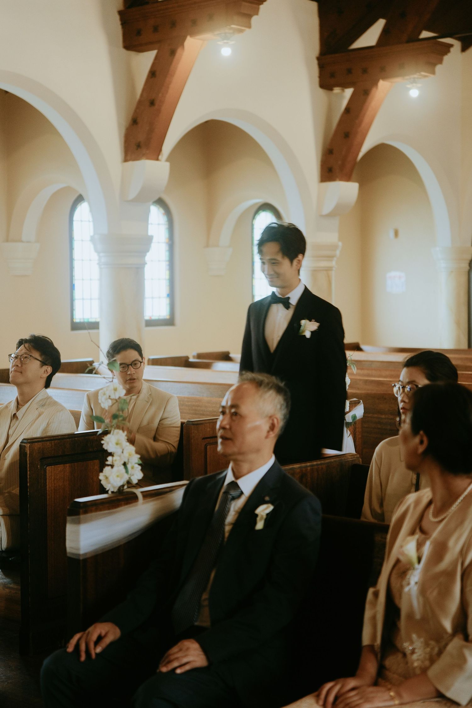 Groom in black tuxedo standing in church aisle with seated wedding guests in wooden pews.