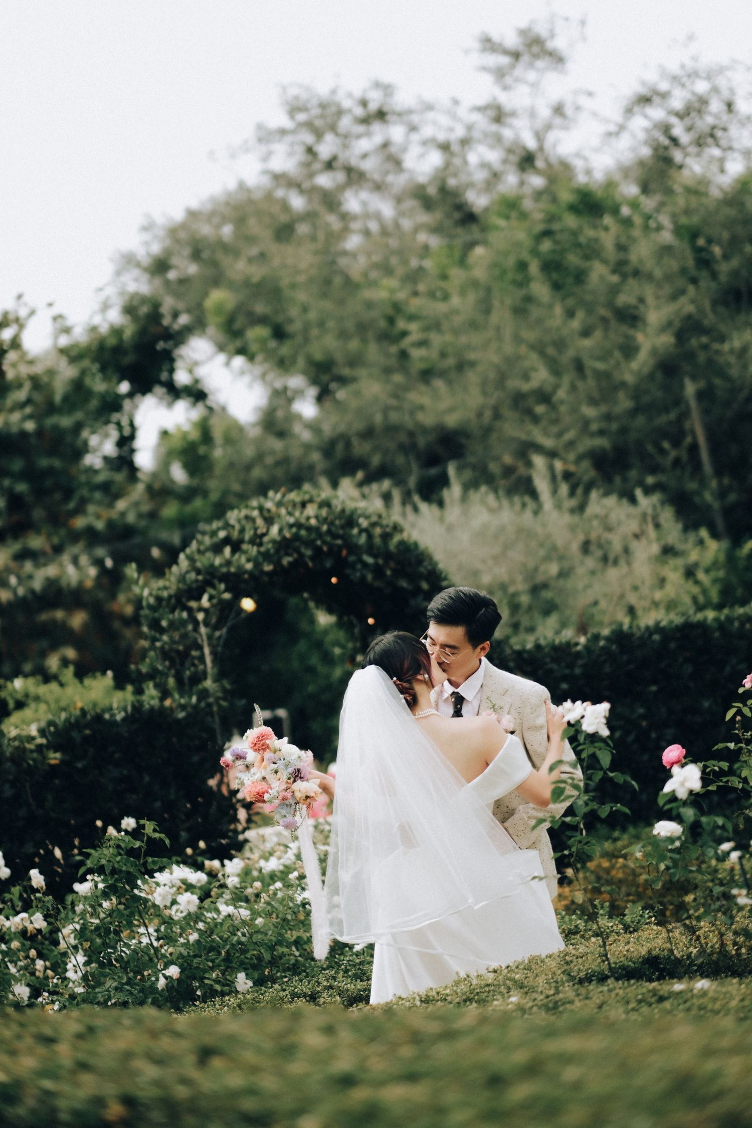 Groom lifting bride in white gown and veil among blooming roses in romantic garden setting.
