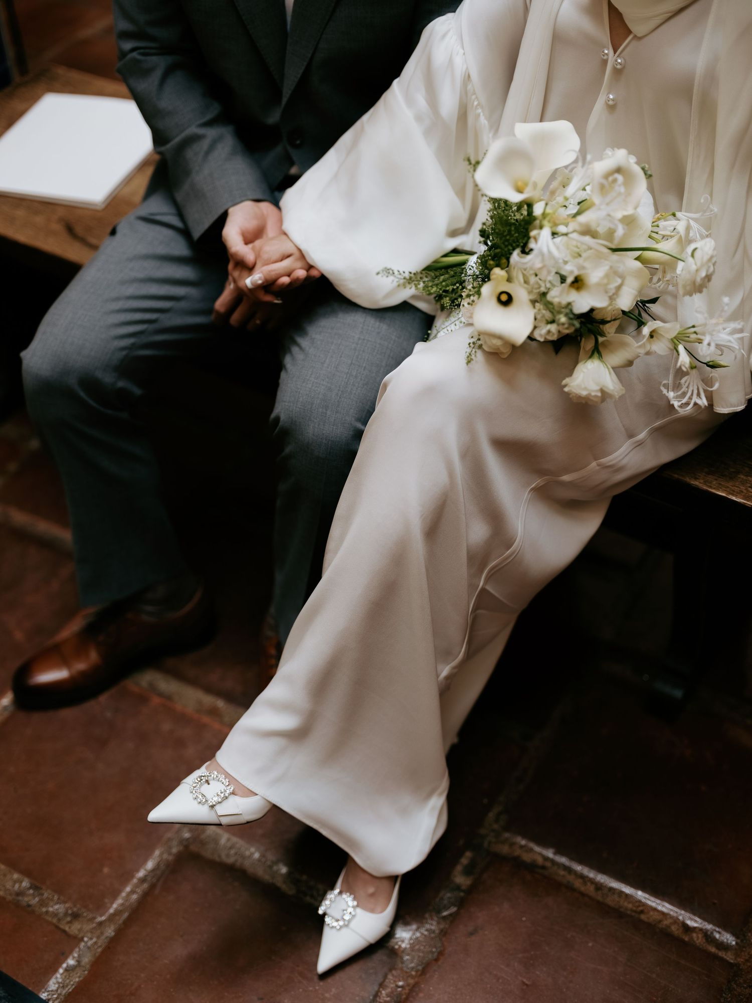 Bride in white satin gown holding floral bouquet holds hands with groom on bench.