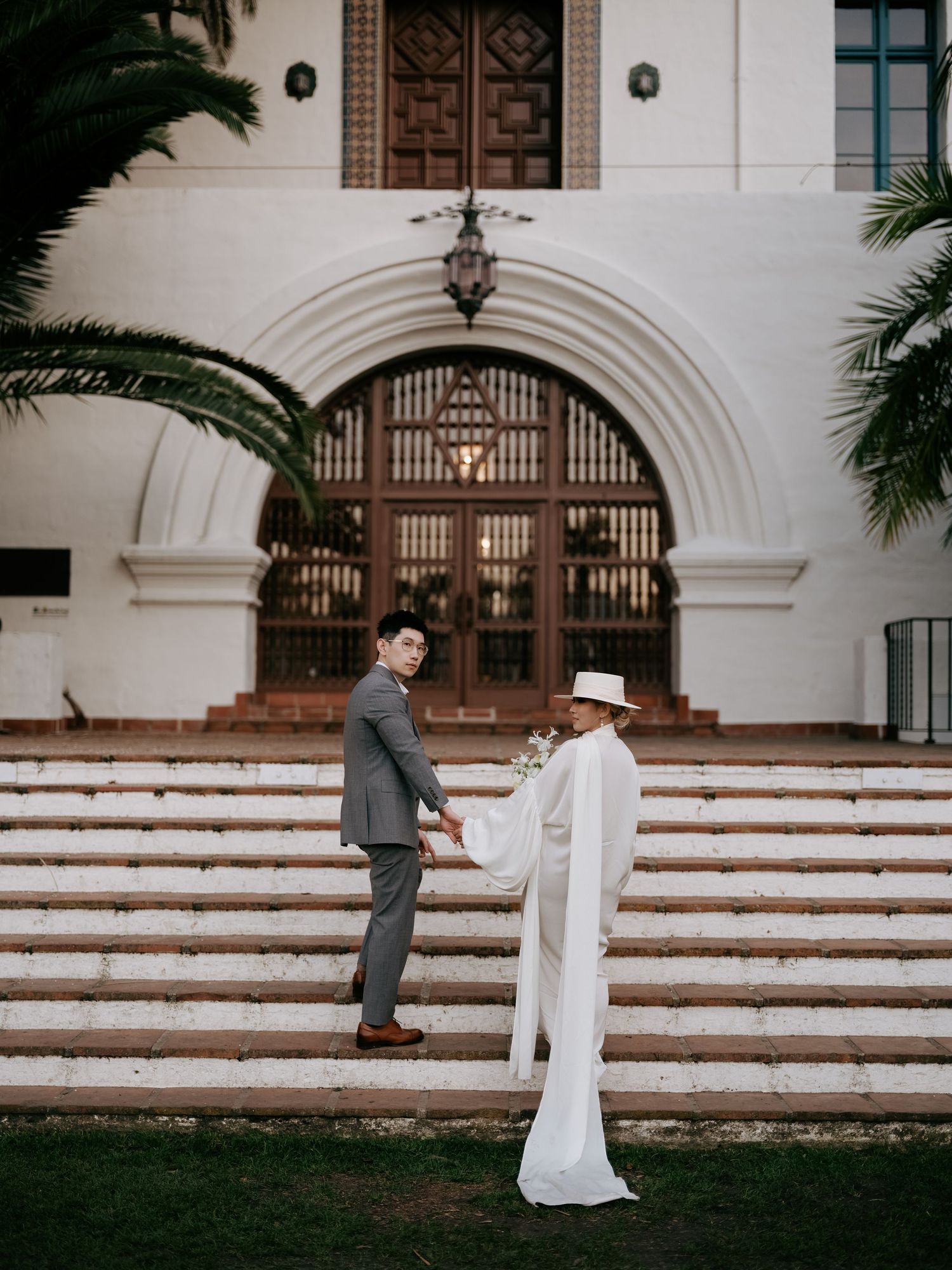 Couple holds hands on steps before grand arched building entrance with palm trees.