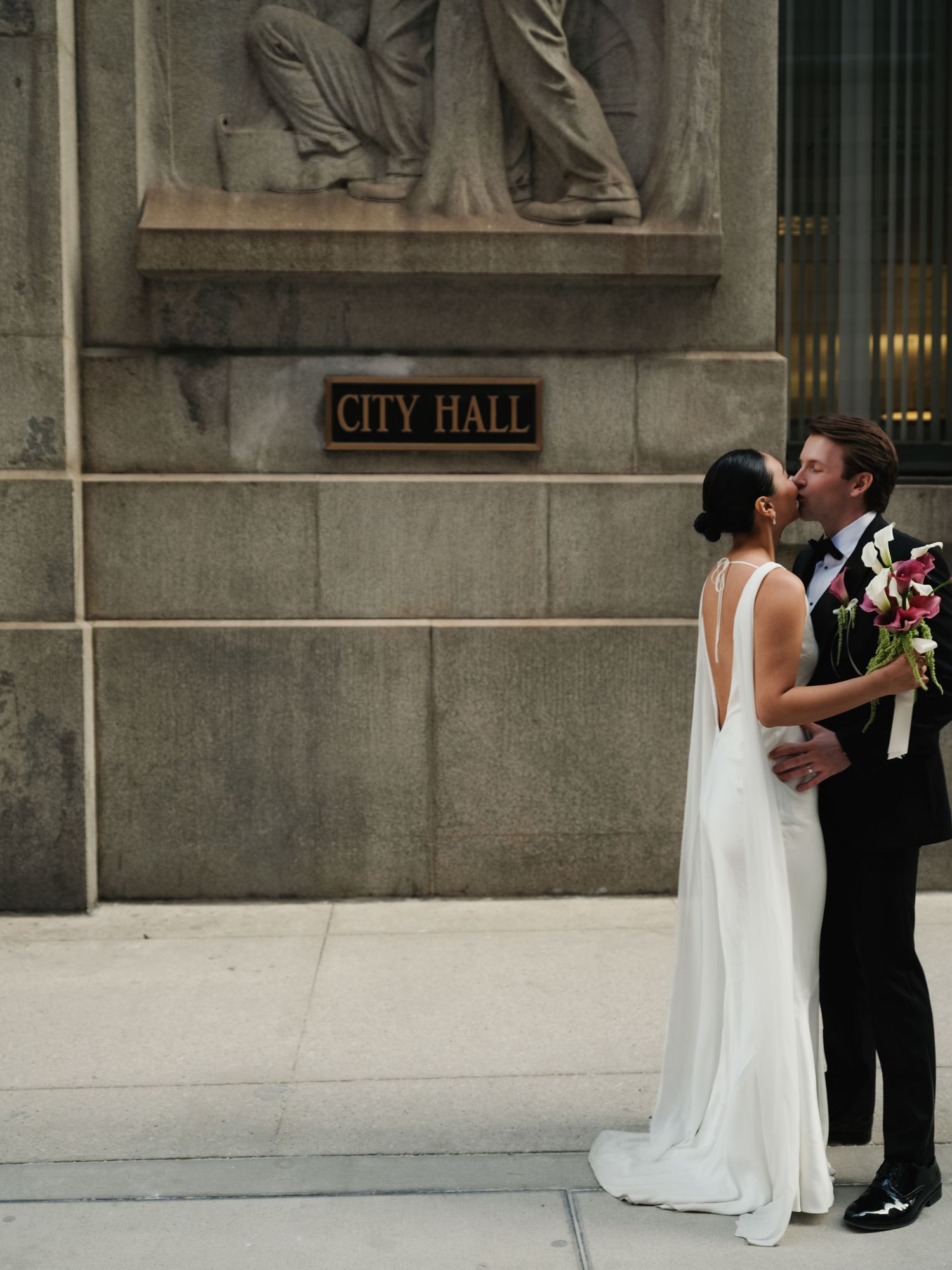Newlyweds sharing a romantic kiss outside City Hall, bride in backless white gown holding colorful bouquet.