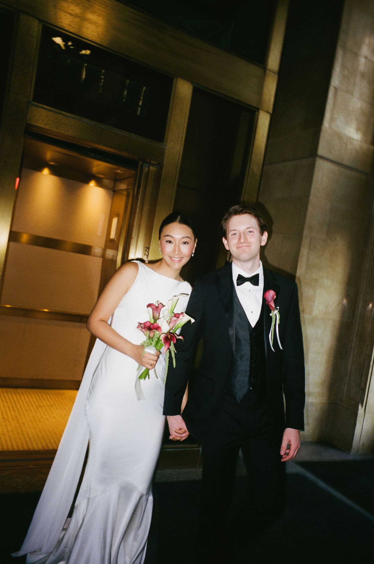 Smiling newlyweds pose in dimly lit corridor, bride holding red calla lily bouquet in white gown.