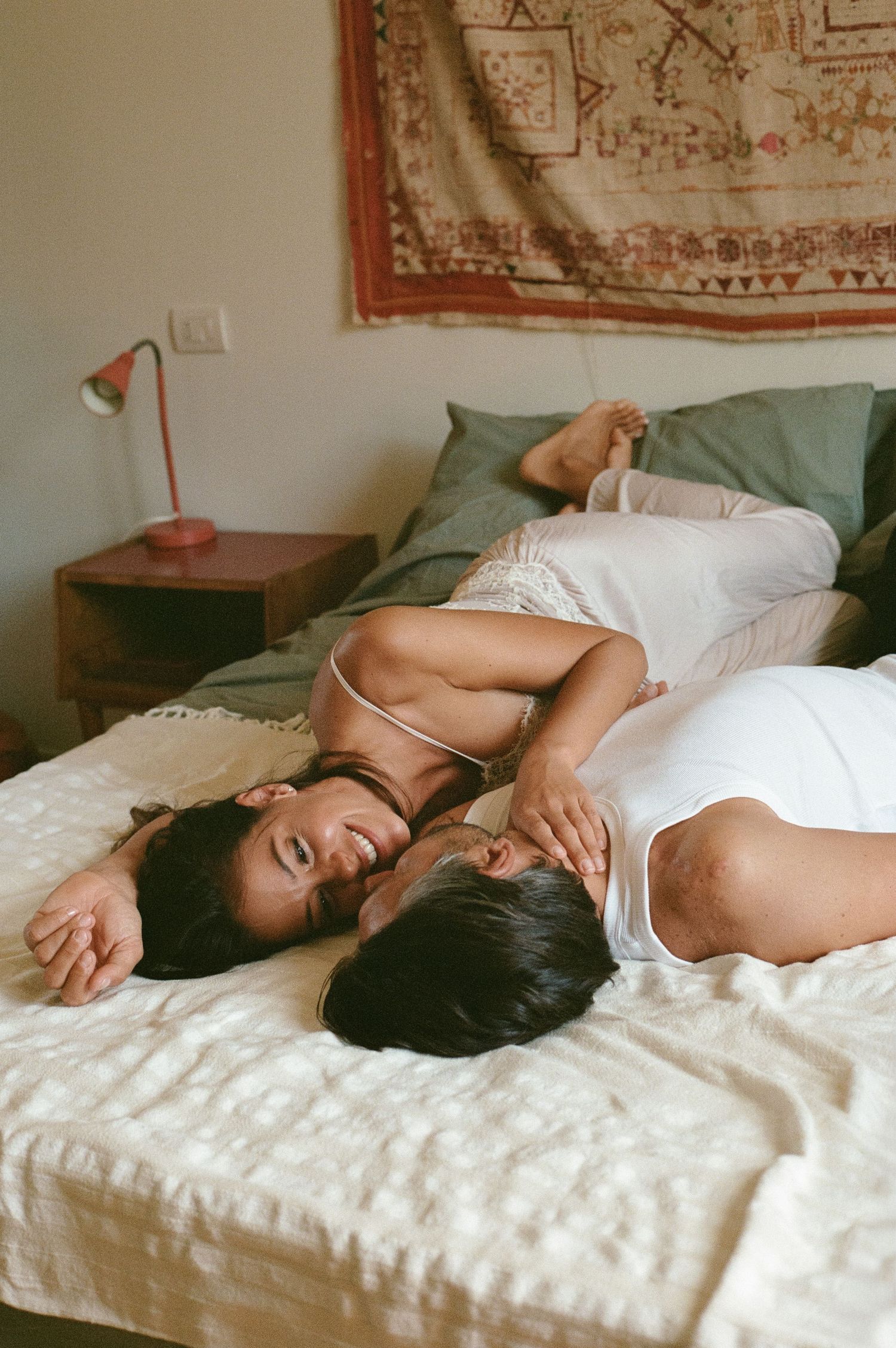 Two women relaxing together on a bed with a bohemian tapestry and red lamp in background.