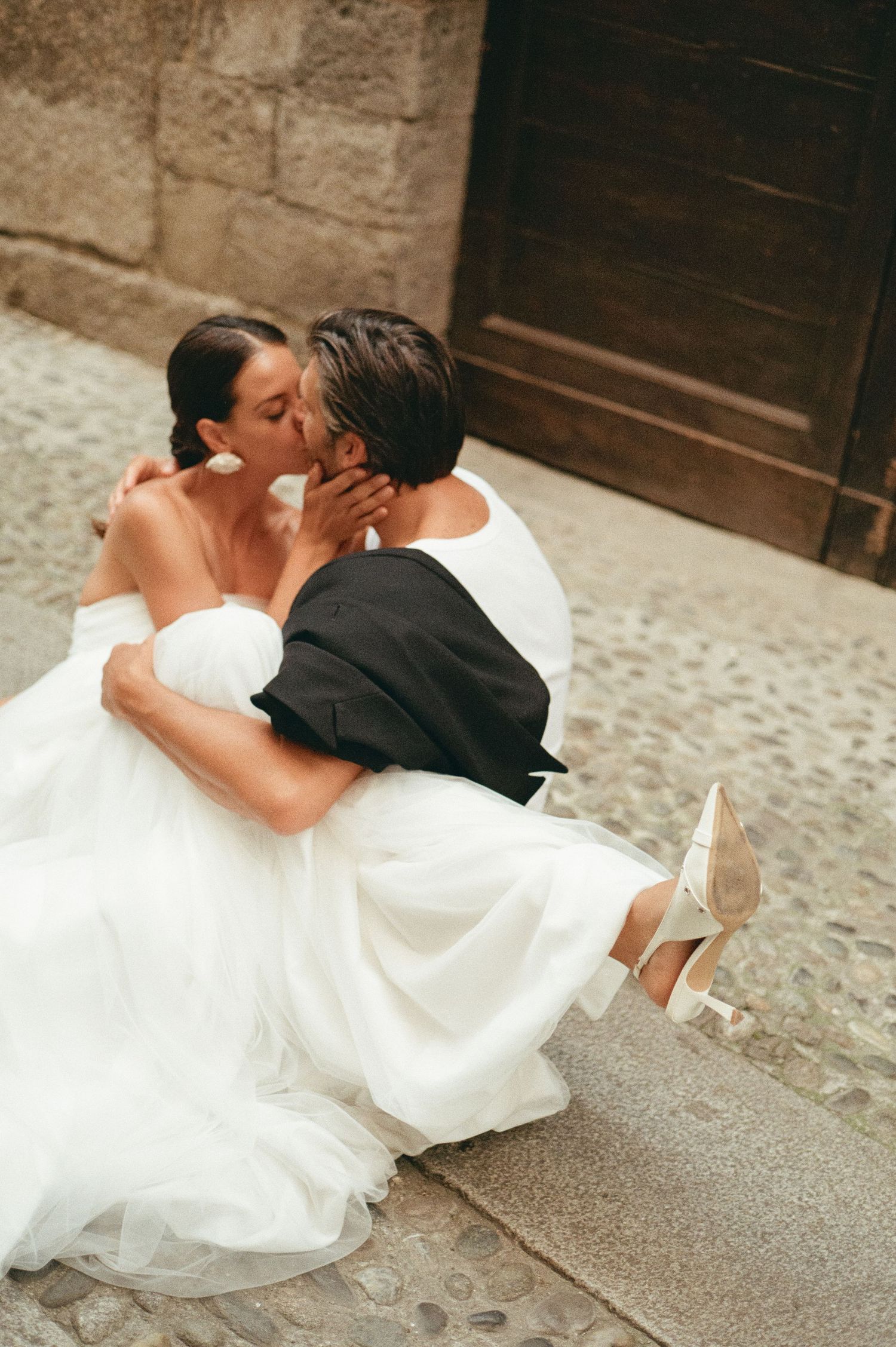 Bride and groom kissing passionately while seated on cobblestone steps beside an old wooden door.