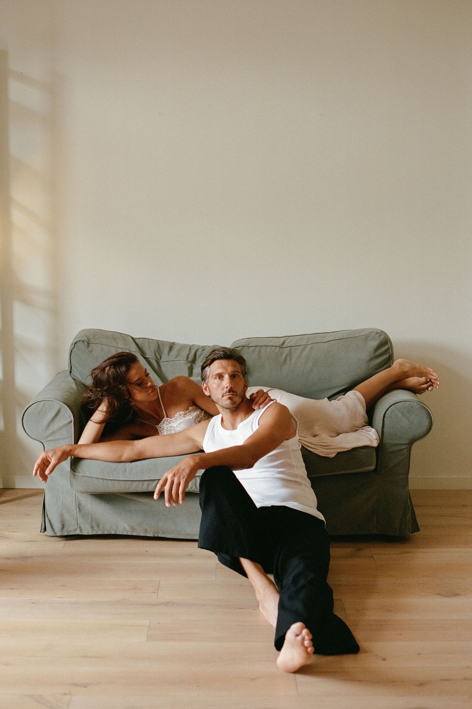 Couple posing casually by a grey sofa, man seated on floor, woman leaning over him.
