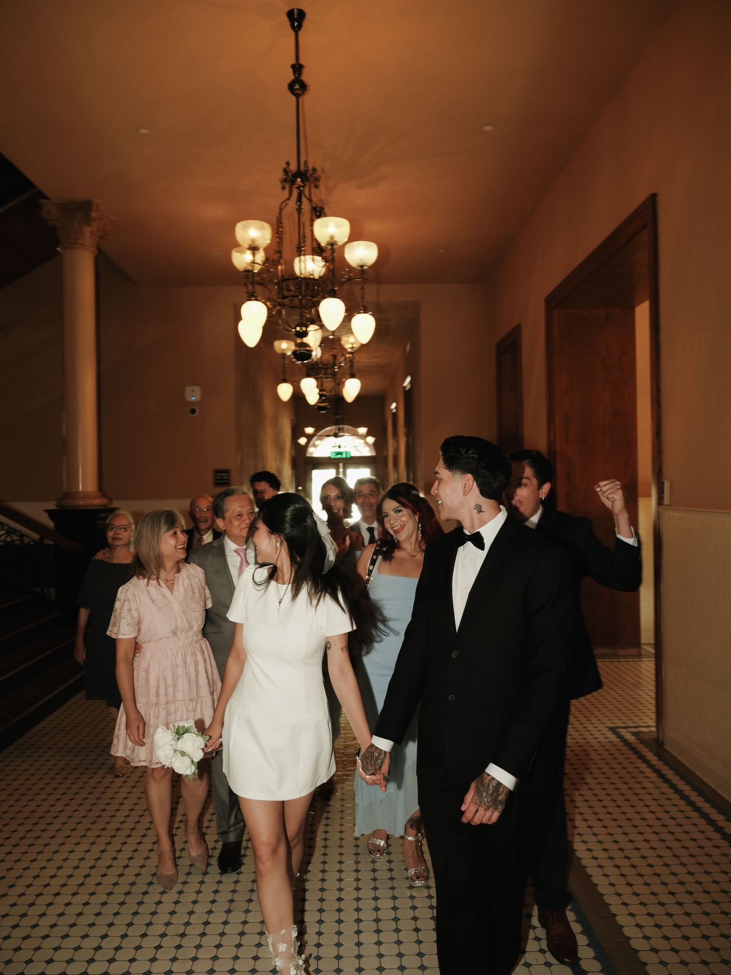 Newlyweds lead wedding party through elegant chandelier-lit hallway, guests celebrating joyfully behind them.