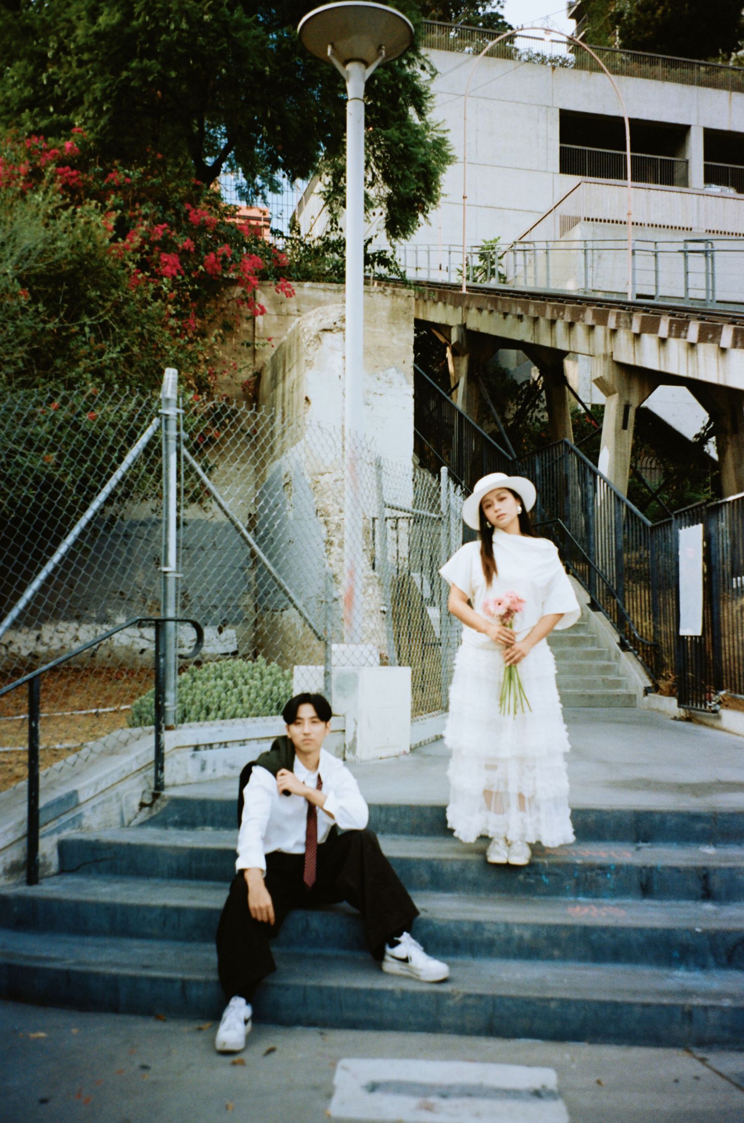 Young couple on urban staircase, woman in white tiered dress and hat with bouquet, man seated on steps below.
