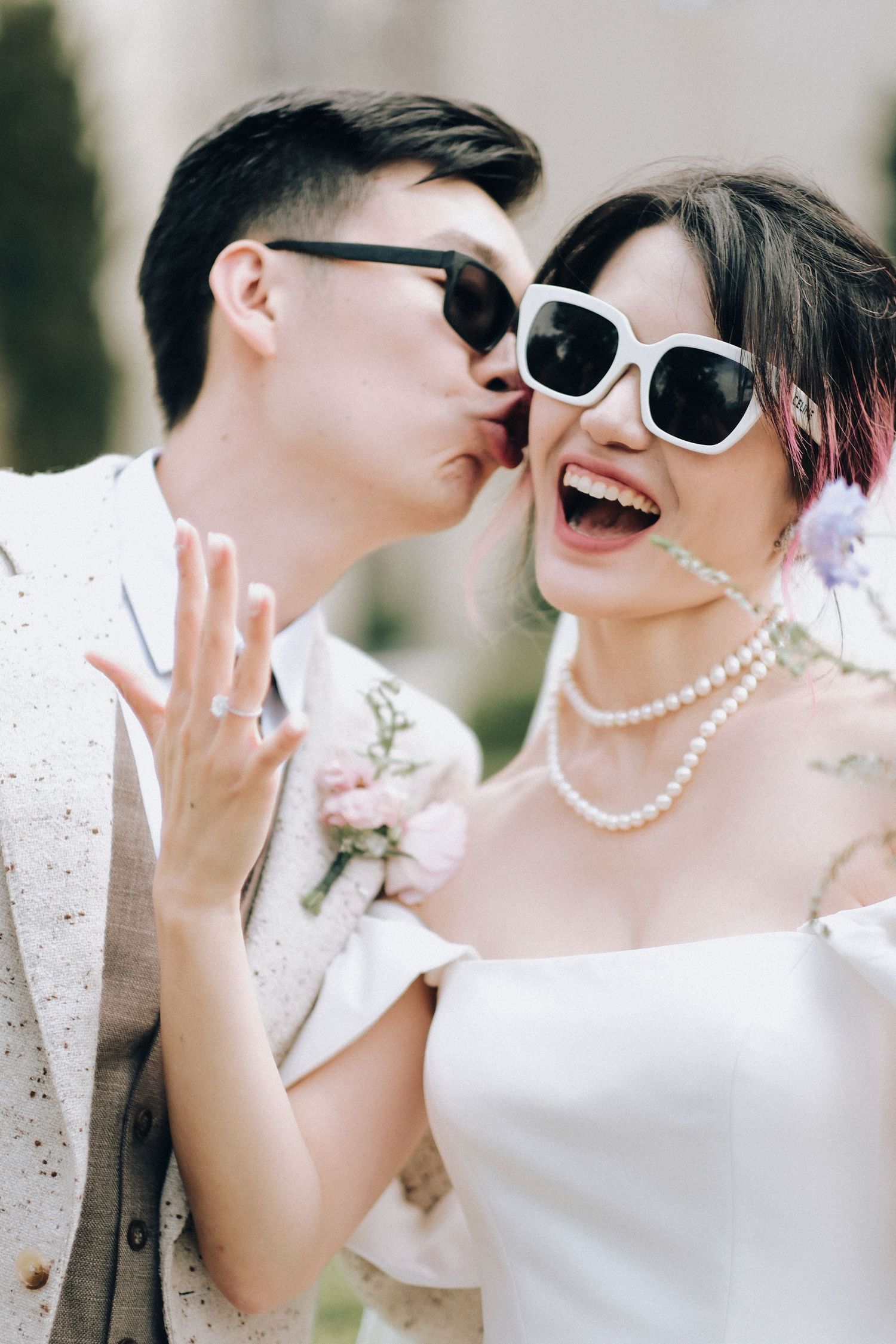Groom kisses laughing bride wearing white sunglasses and pearl necklace at wedding celebration.