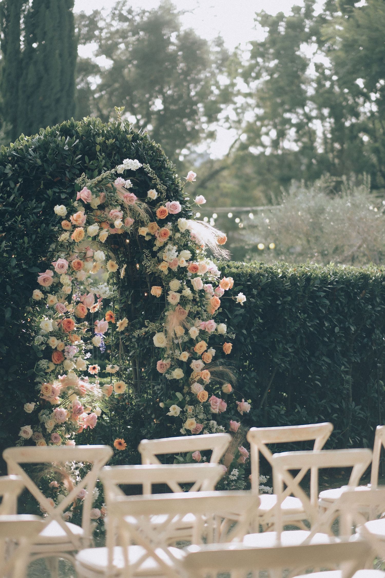Stunning floral wedding arch adorned with pink and peach roses beside manicured hedges and white crossback chairs.