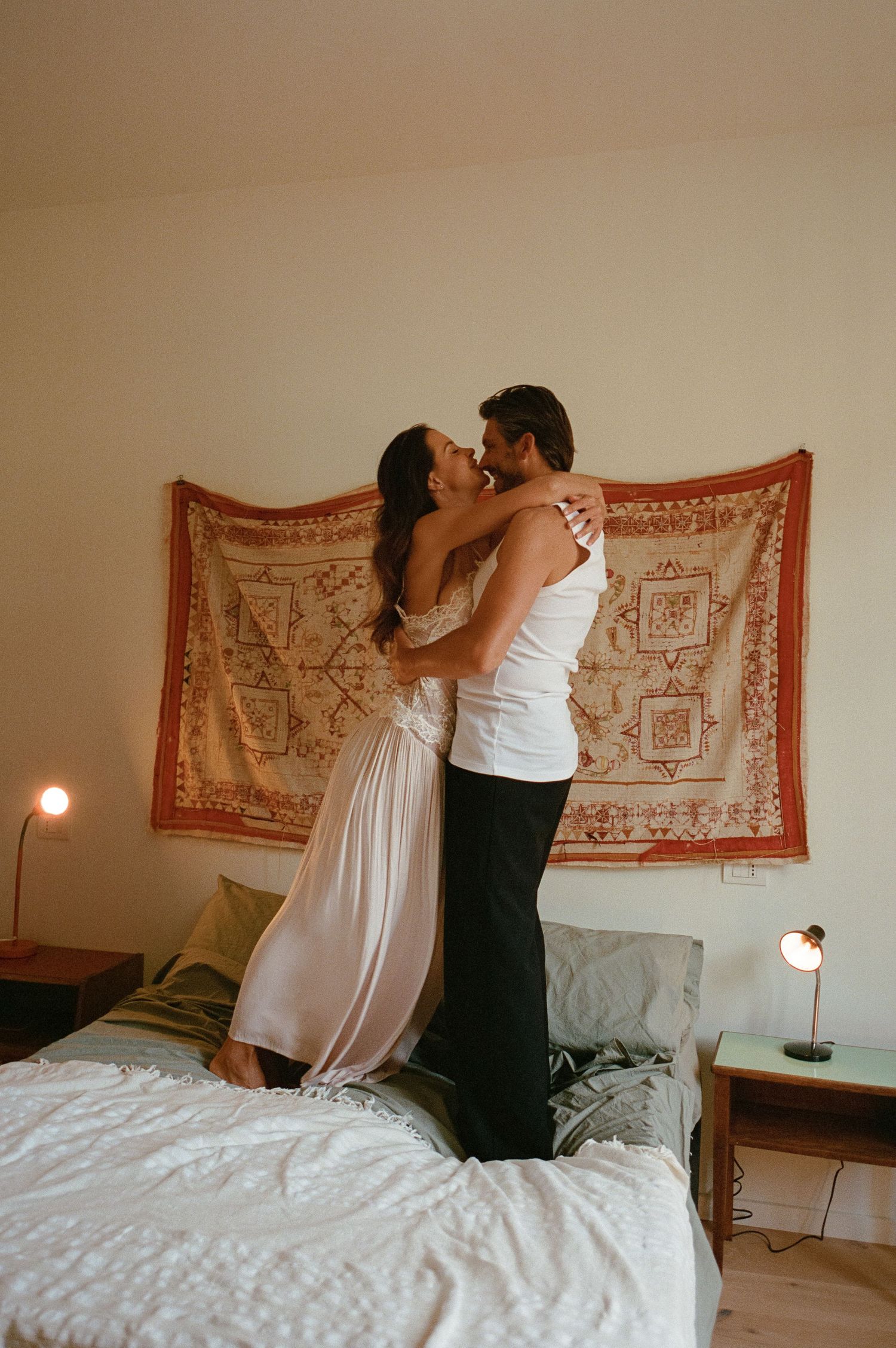 A romantic couple embraces while standing on a white bed, smiling and hugging before a bohemian tapestry backdrop.