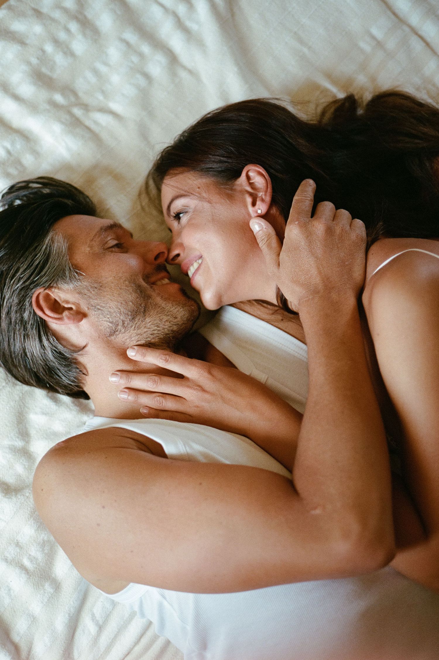 Romantic couple lying on white bed linens smiling and gazing at each other tenderly.