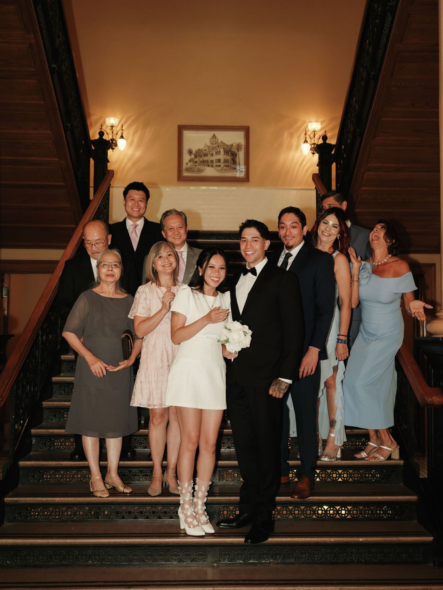 Newlyweds and family gather on elegant staircase for formal wedding portrait in warmly lit venue.
