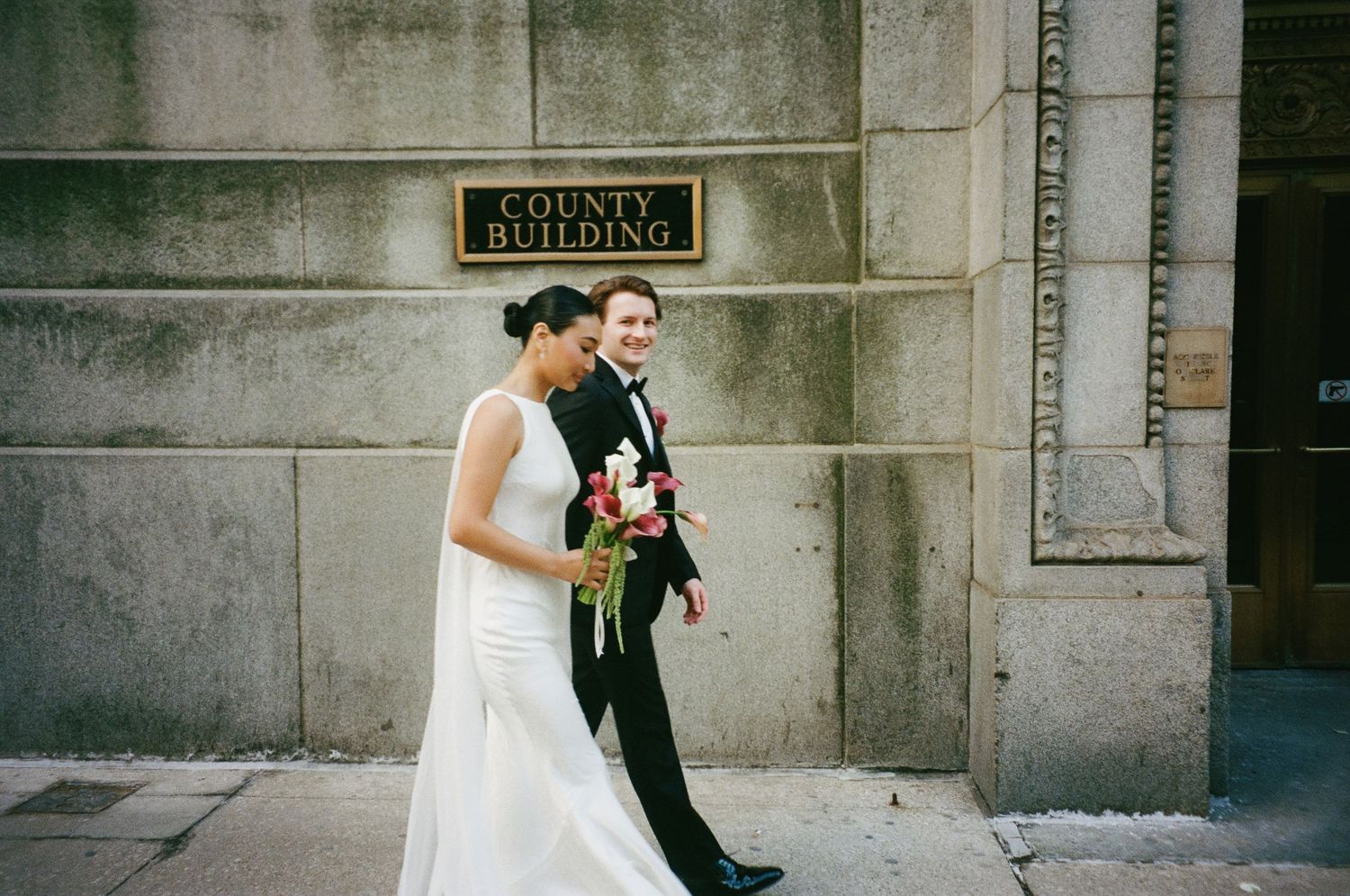 Newlyweds walk past the County Building, bride in elegant white gown carrying a vibrant red bouquet.