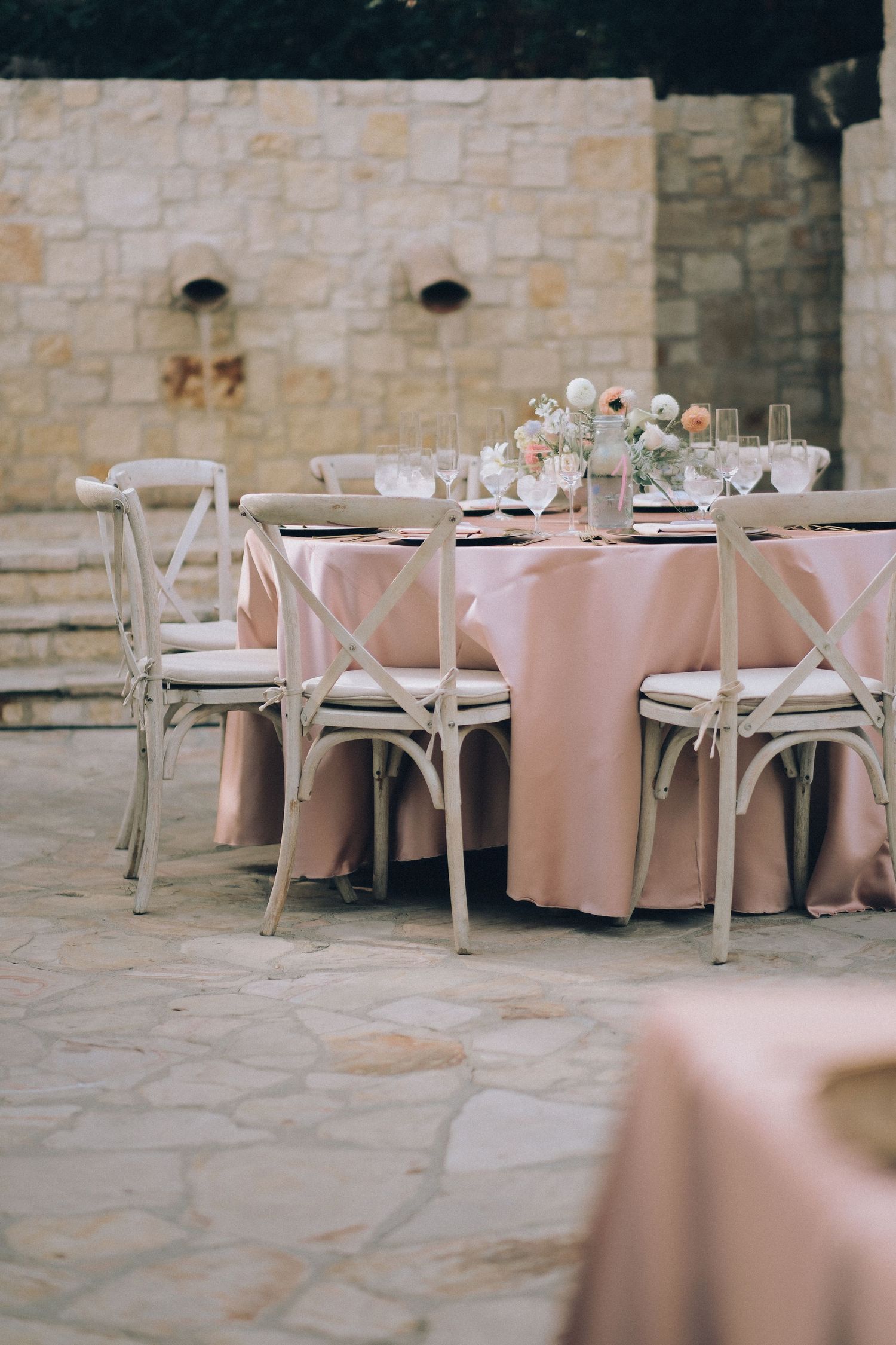 Outdoor wedding reception table with blush pink linen and cross-back chairs set on a stone patio against a stone wall.