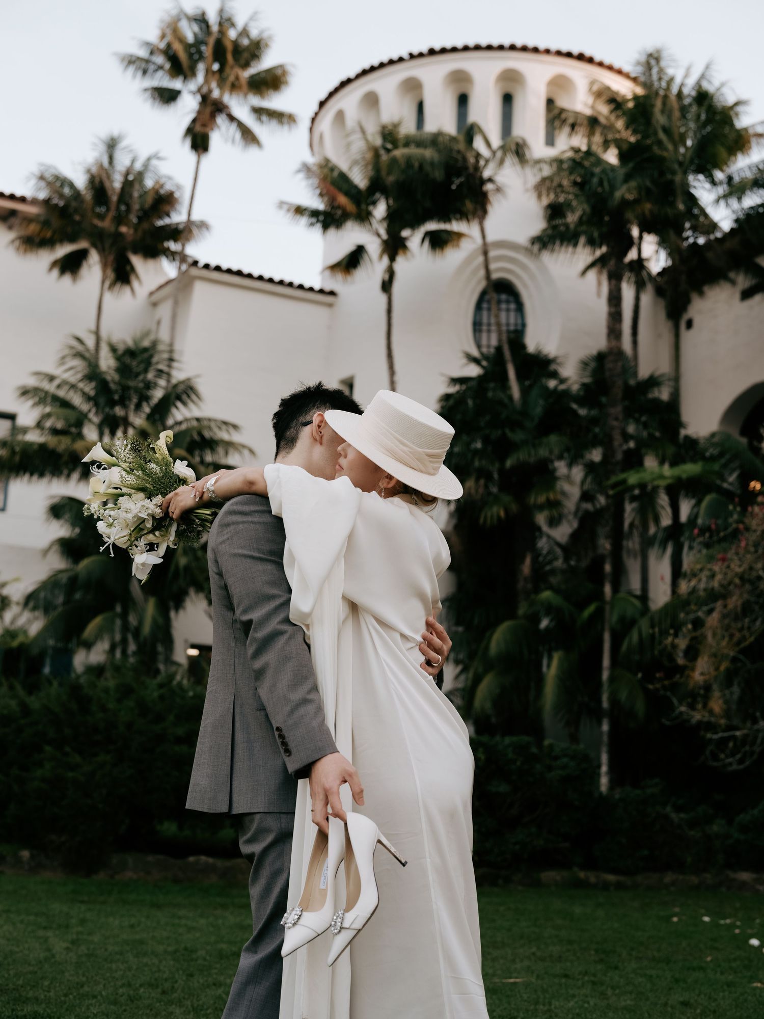 Bride and groom share a romantic kiss outside a Mediterranean-style venue with palm trees.