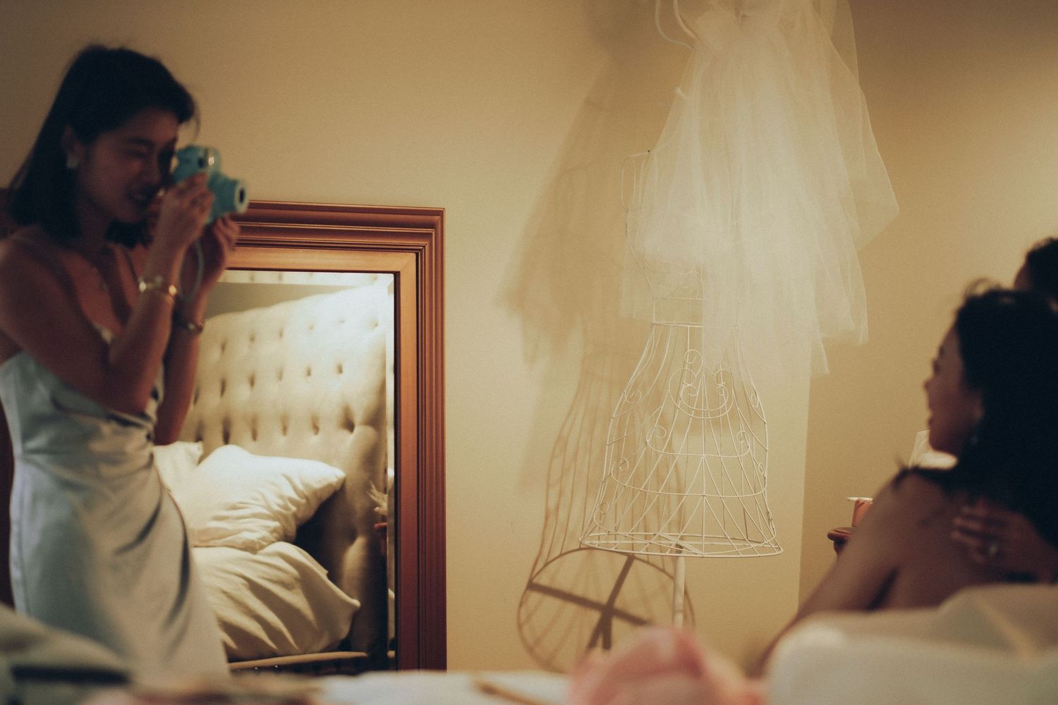 Bridesmaid photographing bride in hotel room with wedding dress hanging on wall before ceremony.