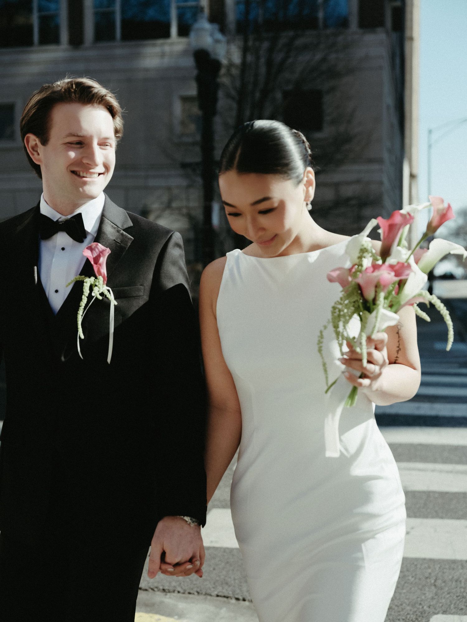 Smiling groom in black tuxedo walks with bride holding pink calla lily bouquet on sunny city street.