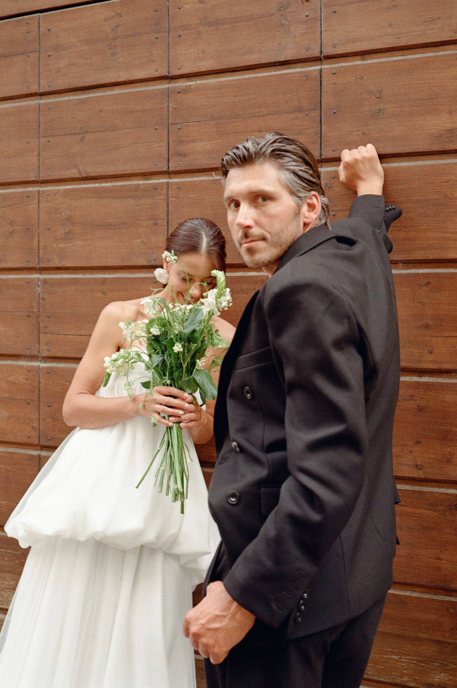 Groom in black suit stands beside bride holding white flower bouquet against wooden wall.