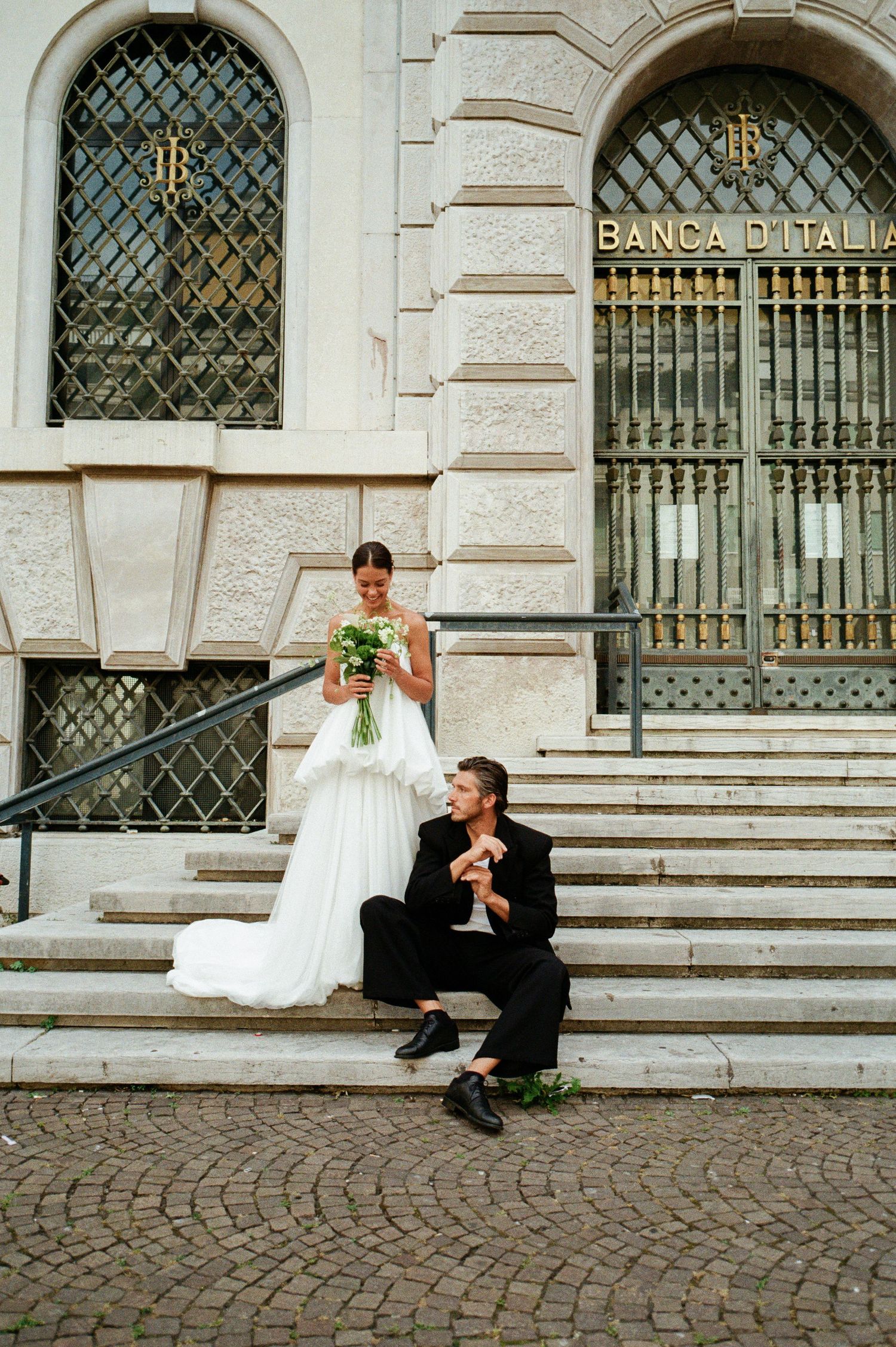 Bride in white ruffled gown and groom sit on steps outside Banca d Italia building in Italy.