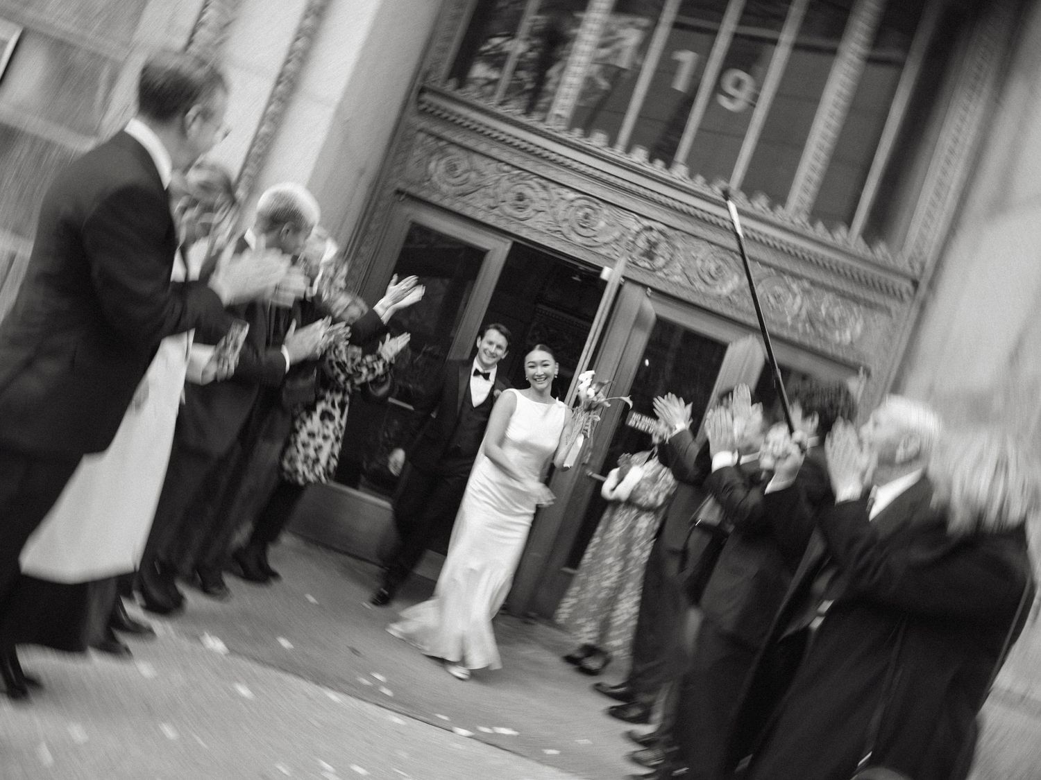 Black and white photo of newlyweds exiting ornate building through cheering wedding guests.