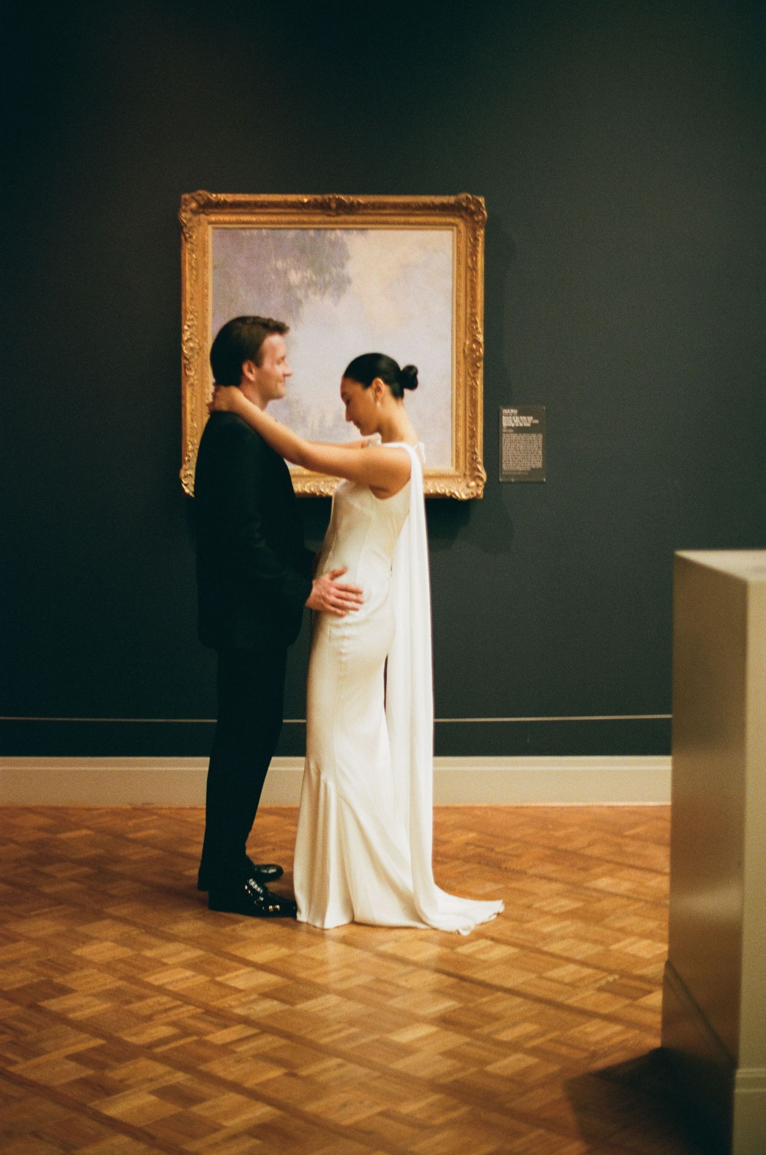 Bride and groom embrace tenderly in front of a large gilt-framed Impressionist painting in a museum gallery.