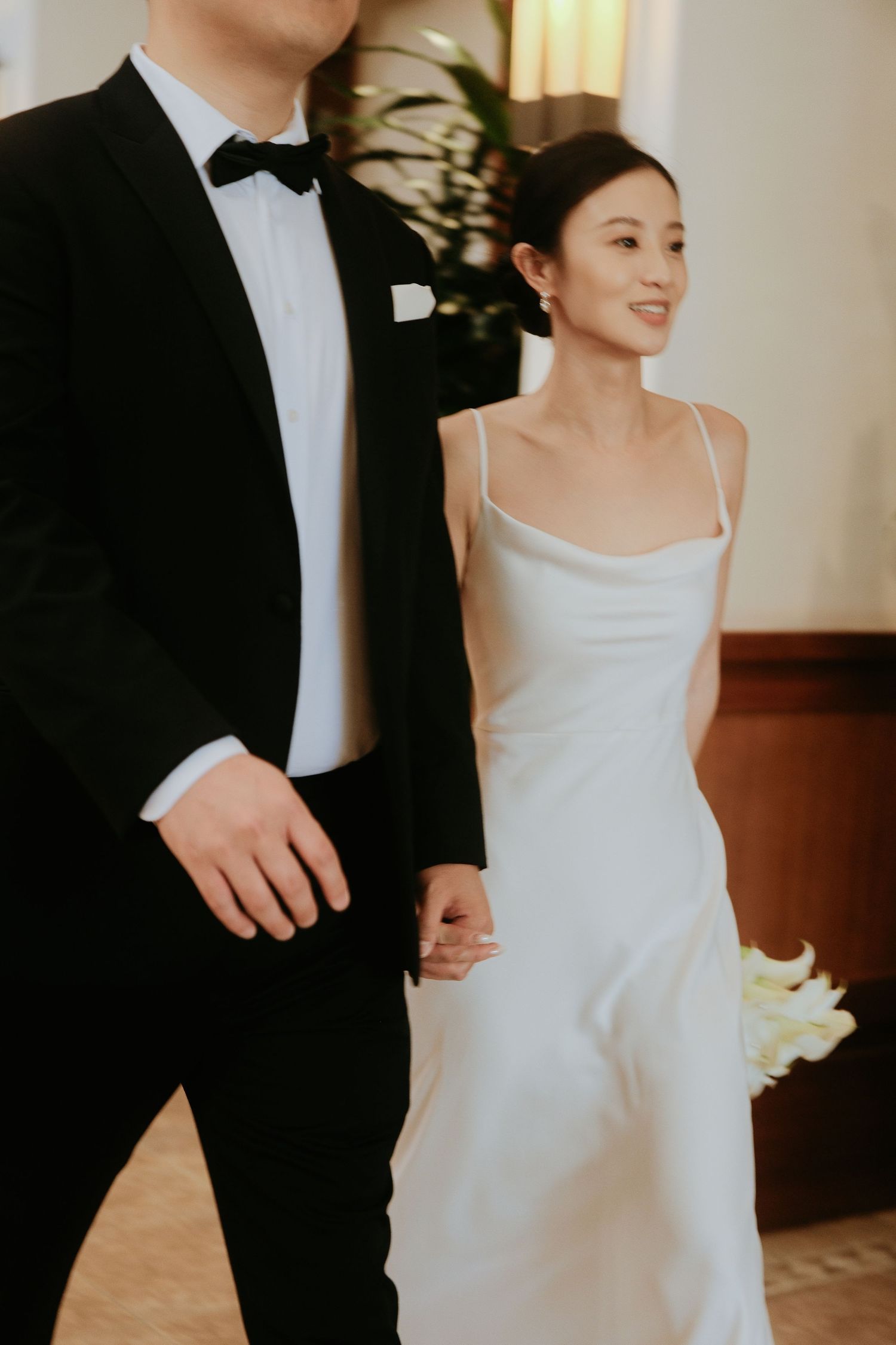 Asian bride in white satin slip dress holds groom's hand while smiling, groom in black tuxedo with bow tie.