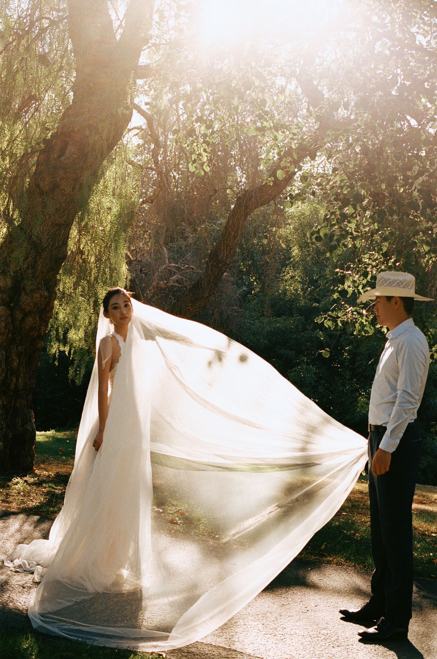 Bride's long white veil billows dramatically in the breeze as groom in cowboy hat watches in sunlit garden.